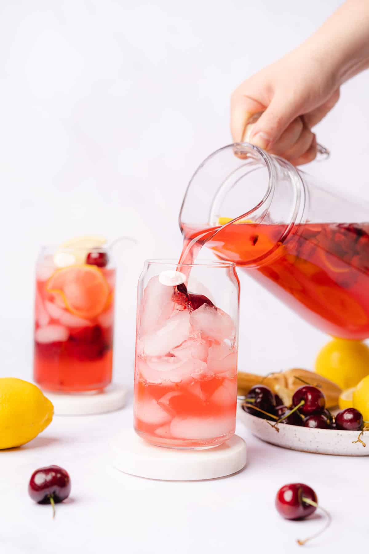 hand pouring from pitcher of cherry lemonade into an ice filled glass