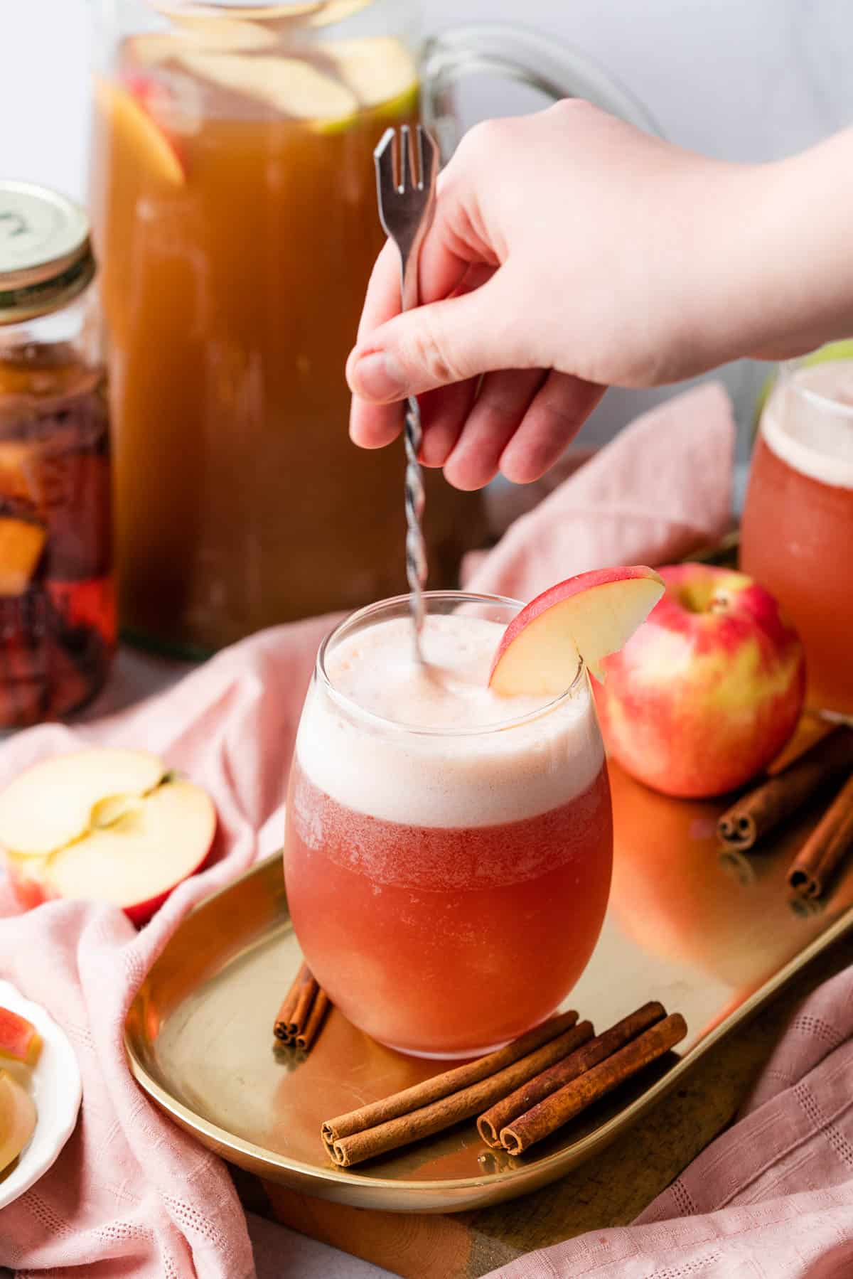 hand stirring a beautiful pink apple cocktail, with apple cider and cinnamon whiskey in the background. 