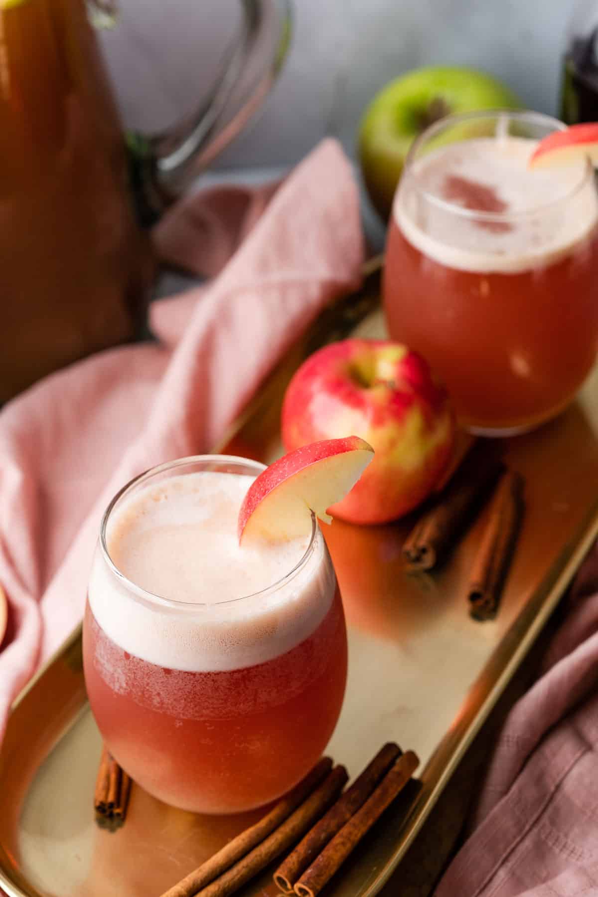 apple cider bourbon cocktails on a gold tray with apples and cinnamon sticks surrounding it  