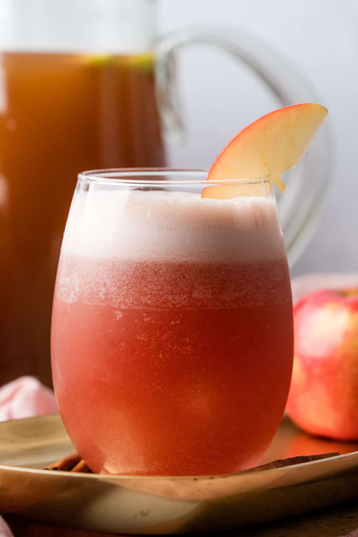 apple cider bourbon cocktail with a pitcher of homemade apple cider in the background topped with an apple wedge