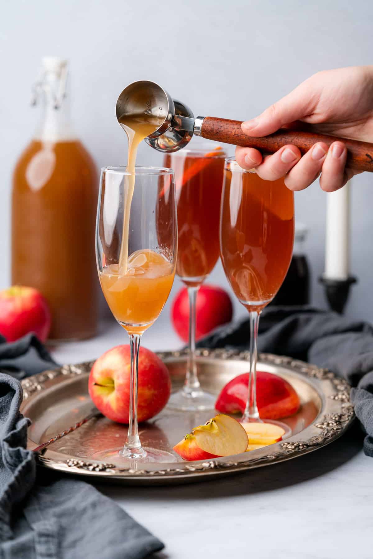 hand pouring apple cider into a champagne flute surrounded by light red, sparkling cocktails and apples