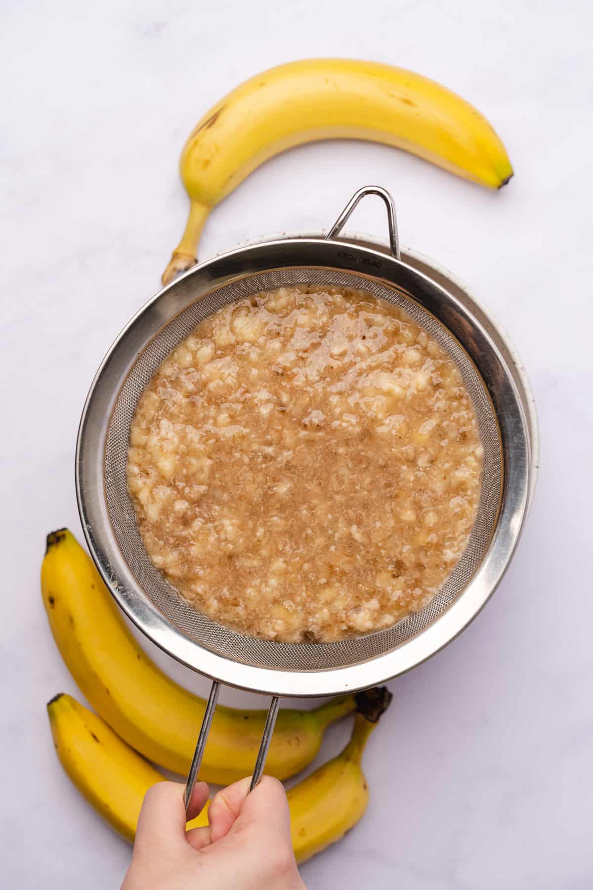 mashed bananas being strained through a metal sieve into a bowl surrounded by fresh bananas