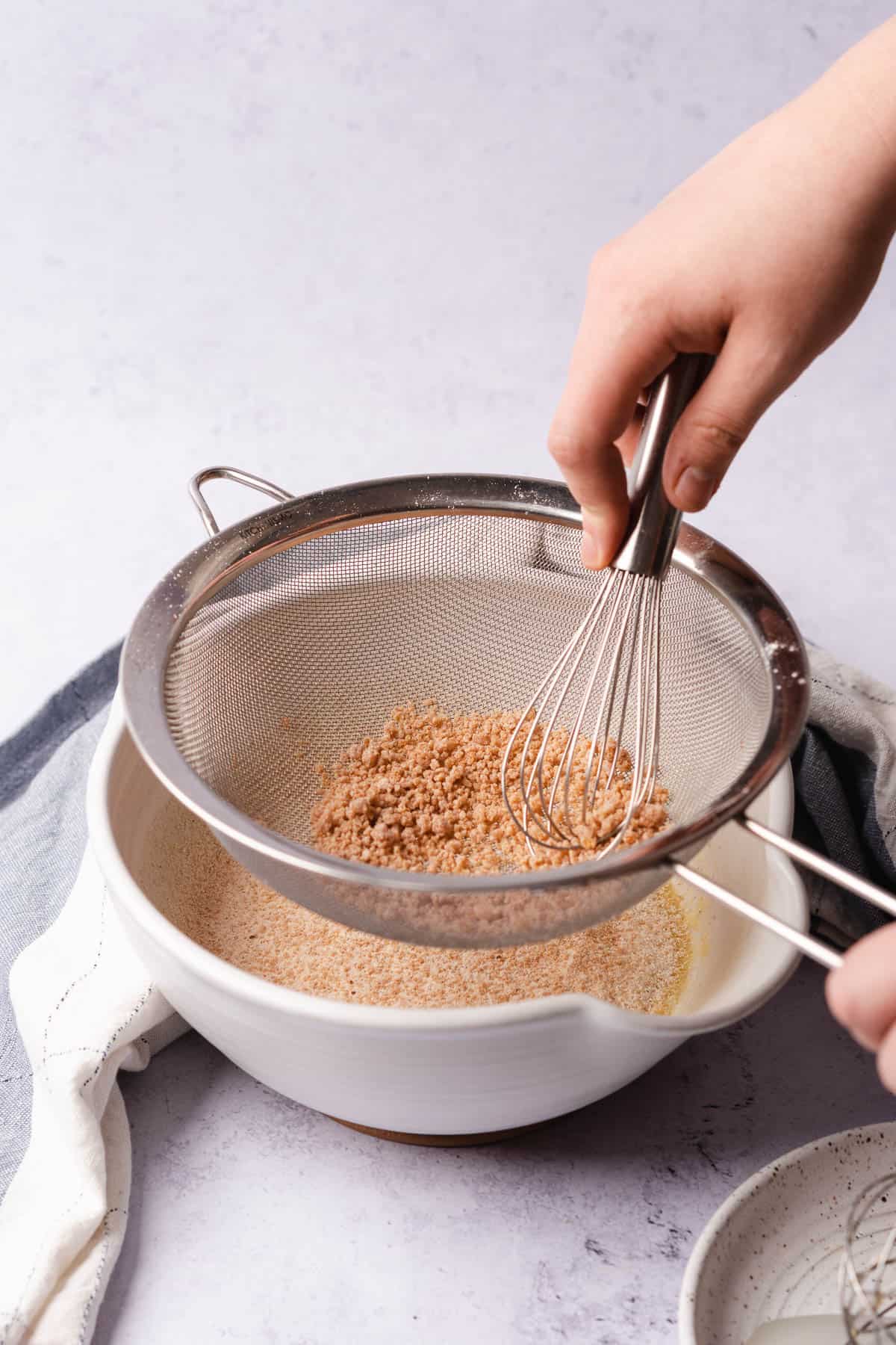 adding dry ingredients to pancake batter by sifting through a metal sieve and pressing with a whisk