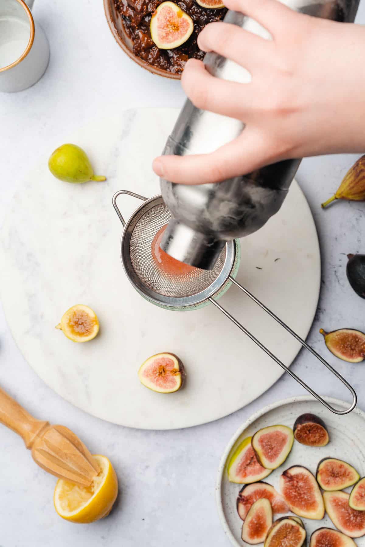 hand pouring fig cocktail contents over a fine mesh strainer