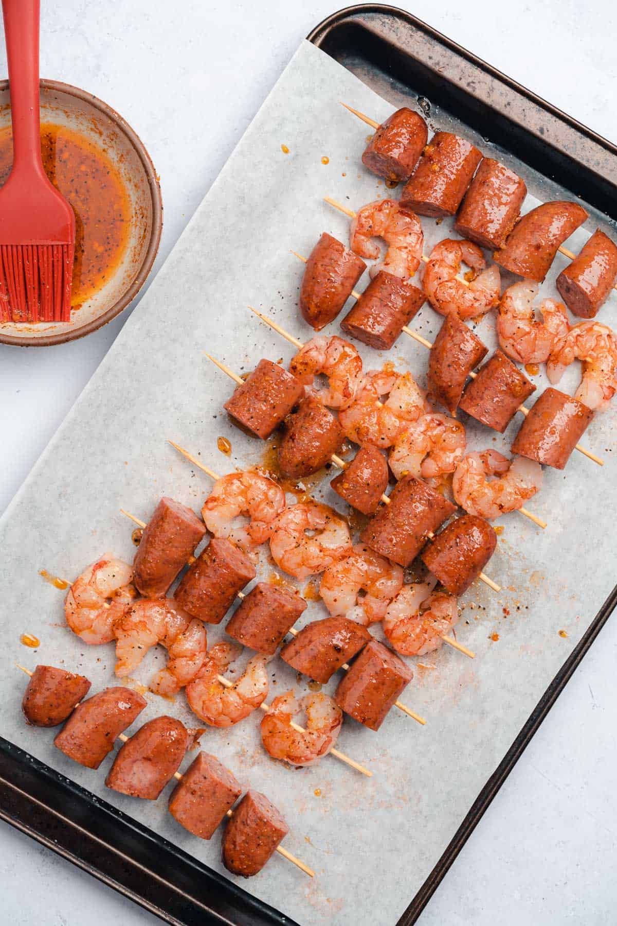 skewered sausage and shrimp on a baking sheet with parchment paper, getting coated in the marinade
