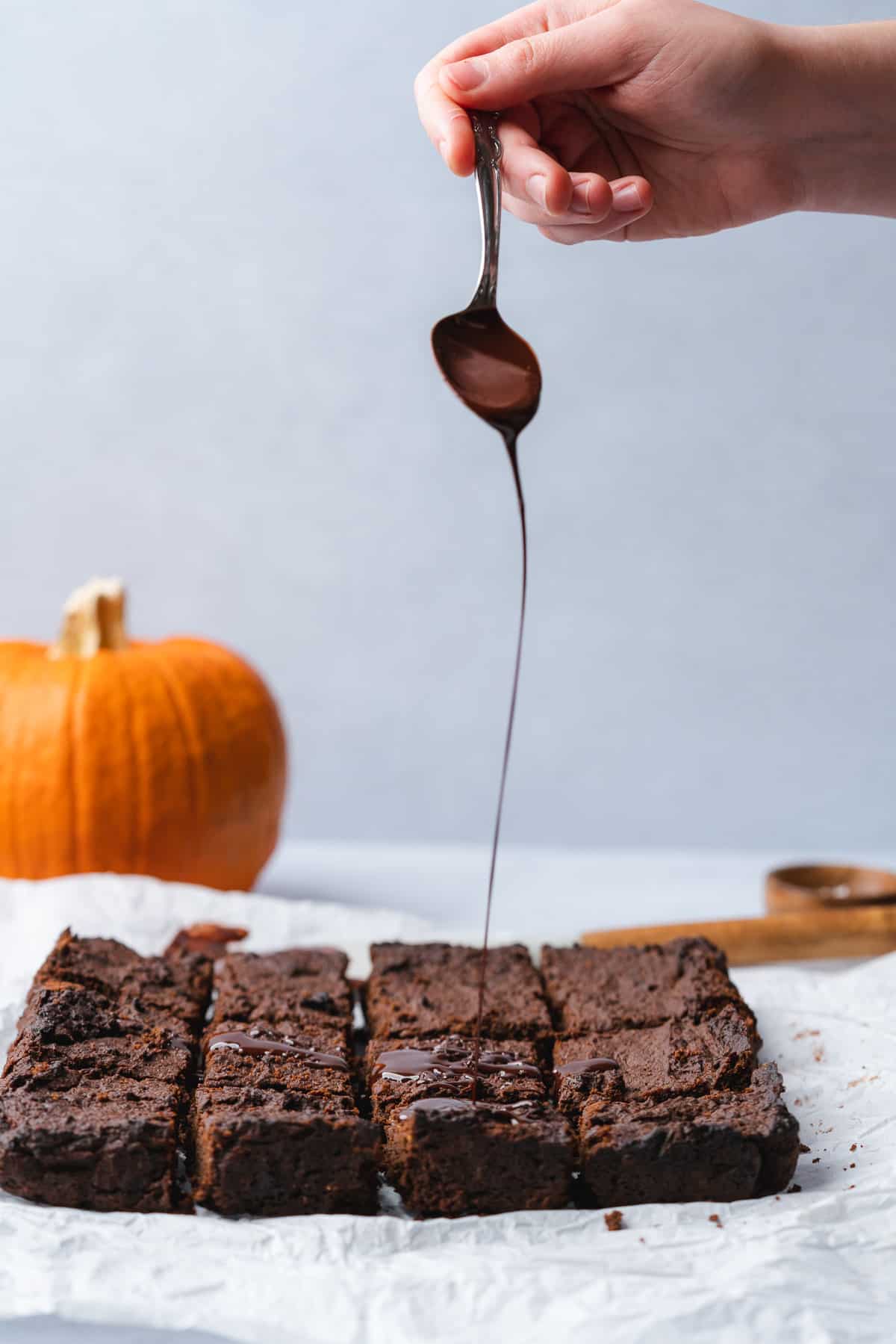 hand drizzling chocolate ganache overtop of the brownies with a pumpkin in the background