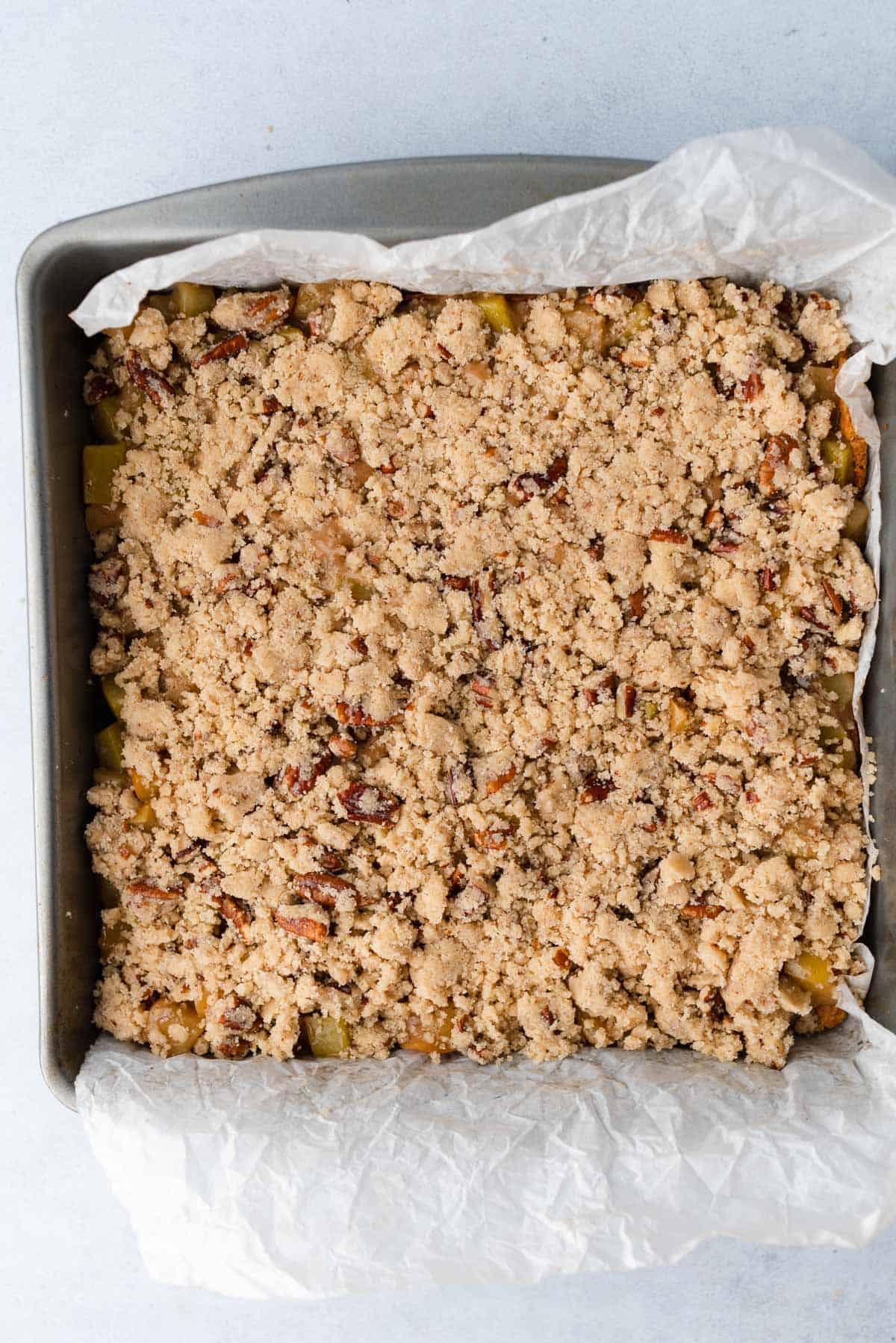 apple pecan crumble topping overtop of apple filling in a baking sheet with parchment