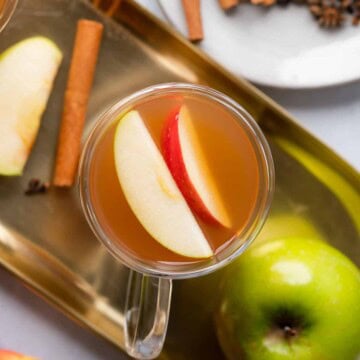 flat lay of homemade low carb apple cider with a granny smith apple and cinnamon sticks inside, alongside fresh apples on a gold tray