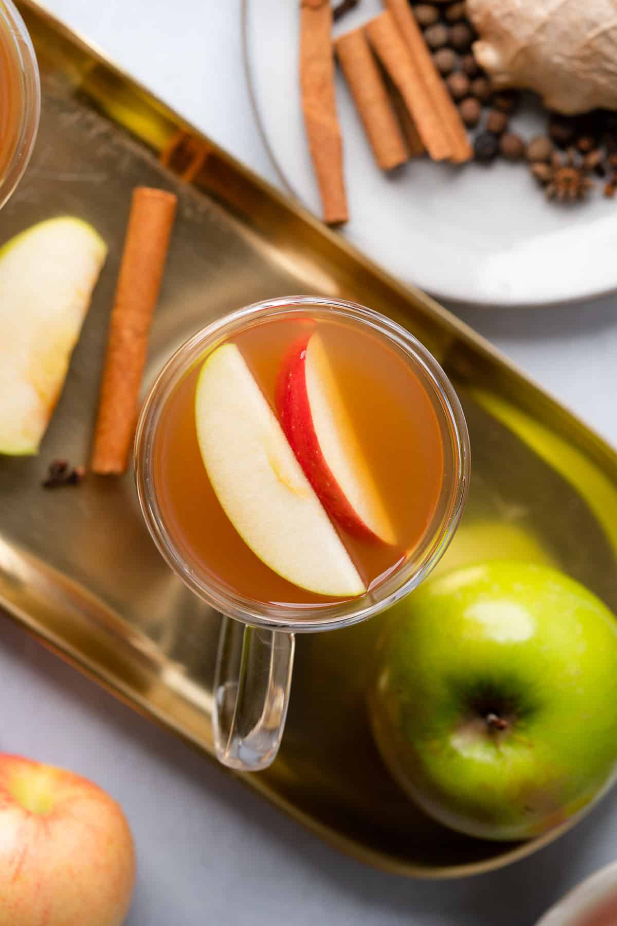 flat lay of homemade low carb apple cider with a granny smith apple and cinnamon sticks inside, alongside fresh apples on a gold tray