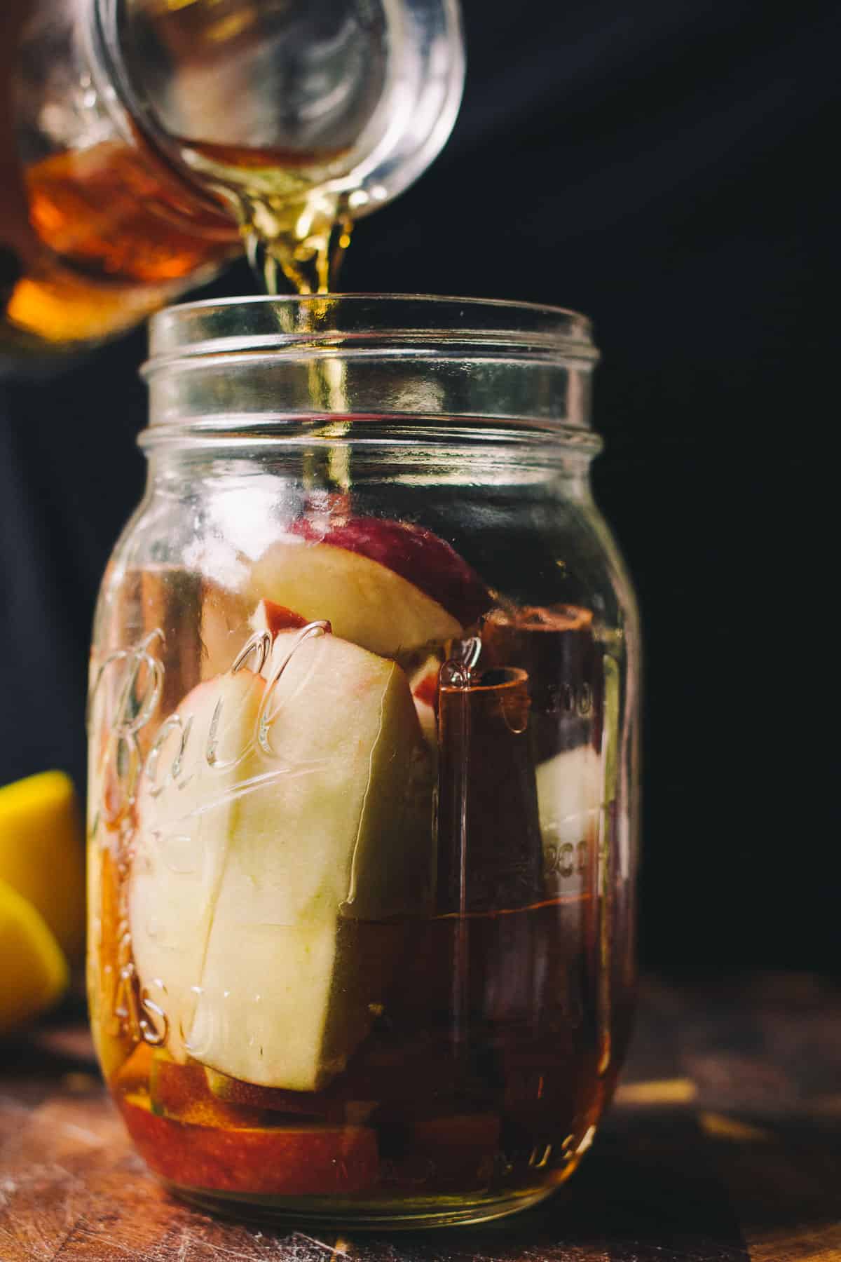 pouring in bourbon whiskey into a glass mason jar with apple slices and cinnamon sticks 