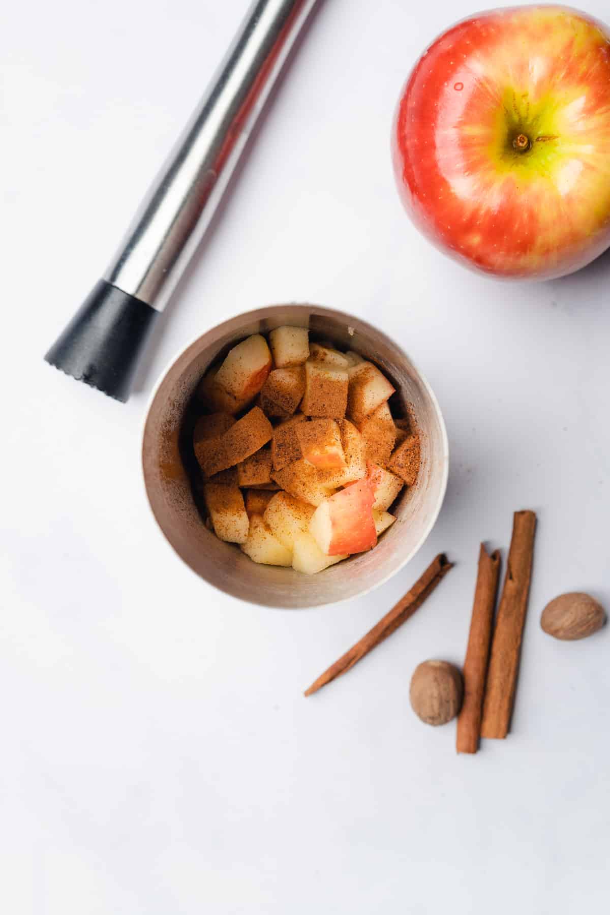 chunks of honey crisp apple  topped with cinnamon and nutmegat the bottom of a cocktail shaker beside a fresh apple, cinnamon sticks, nutmeg and a muddler
