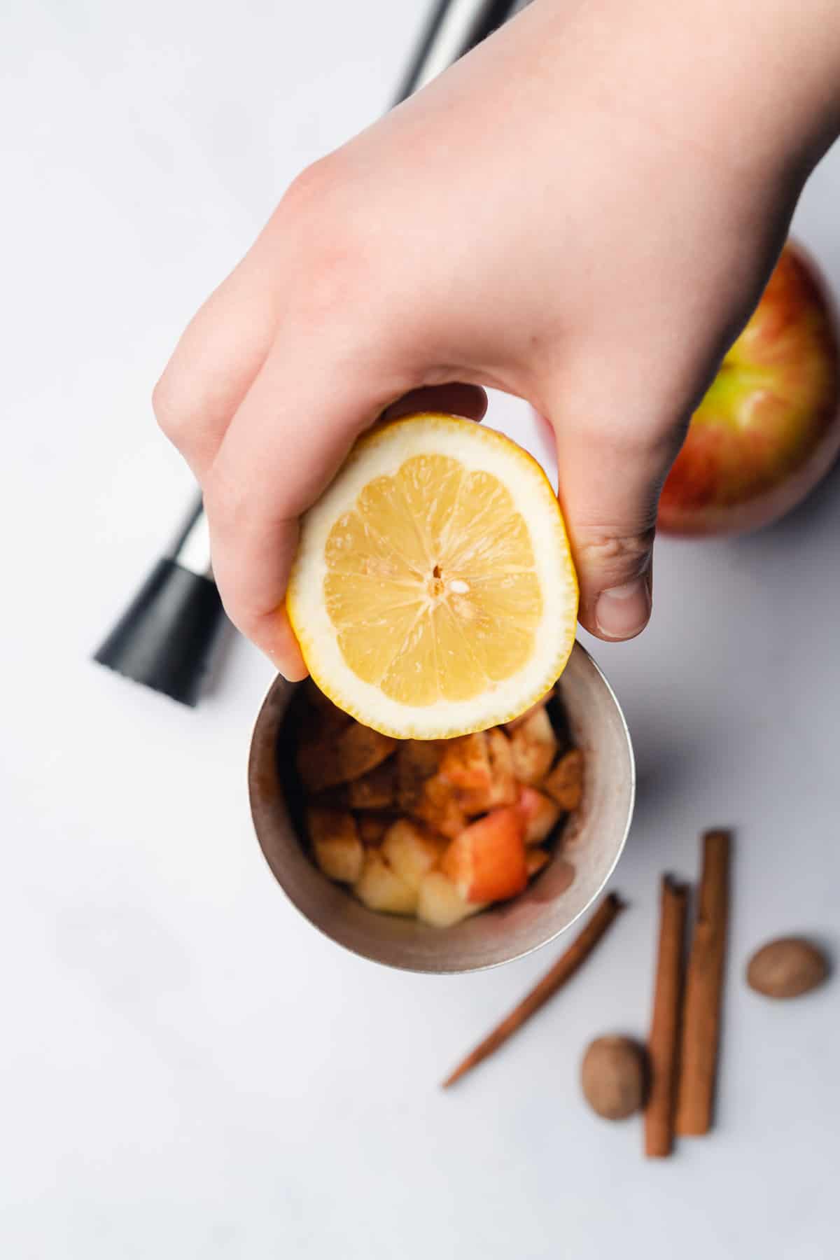 hand holding a lemon over top of a cocktail shaker with honey crisp apple, cinnamon and nutmeg
