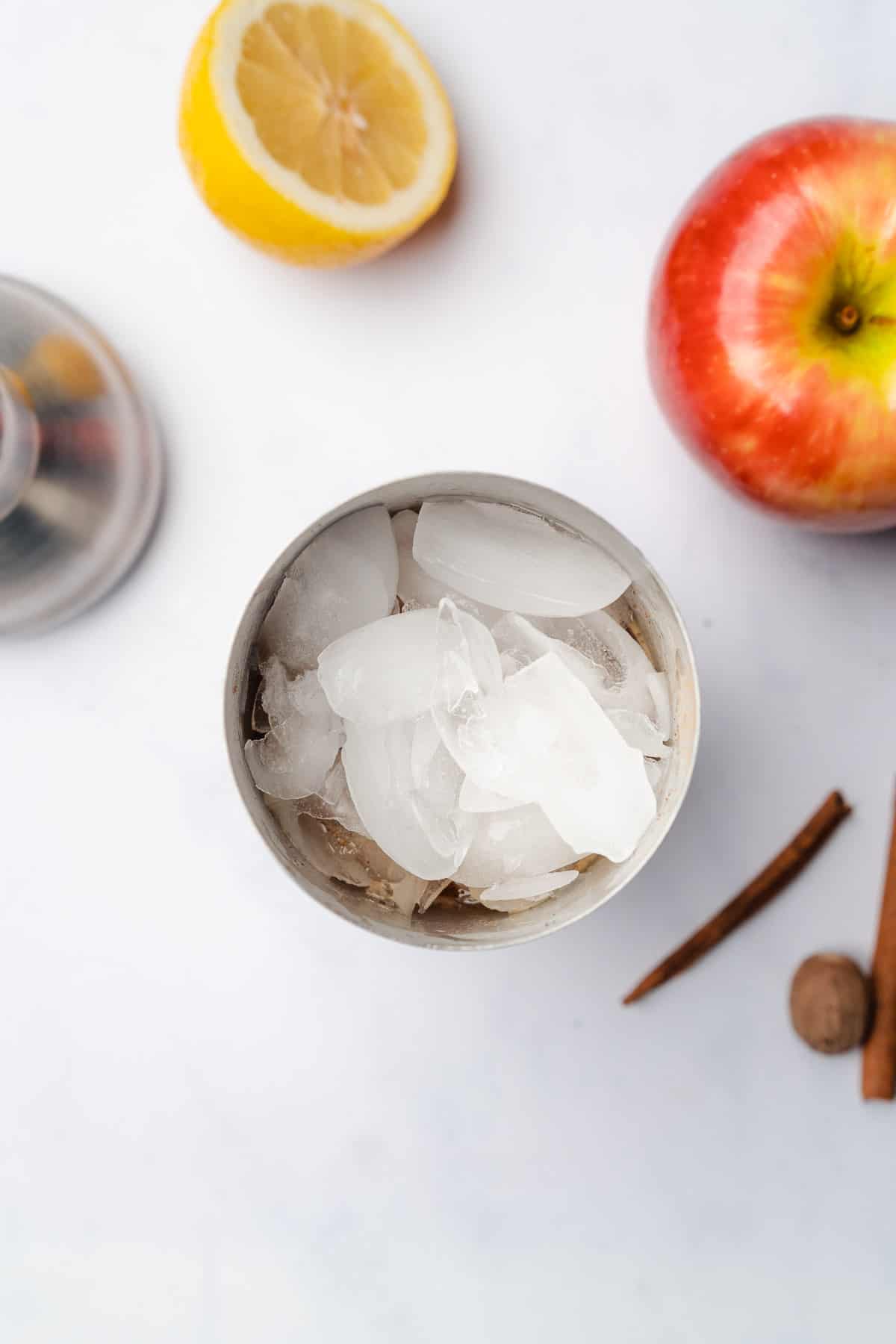 ice in a cocktail shaker surrounded by apples, nutmeg, cinnamon and lemon