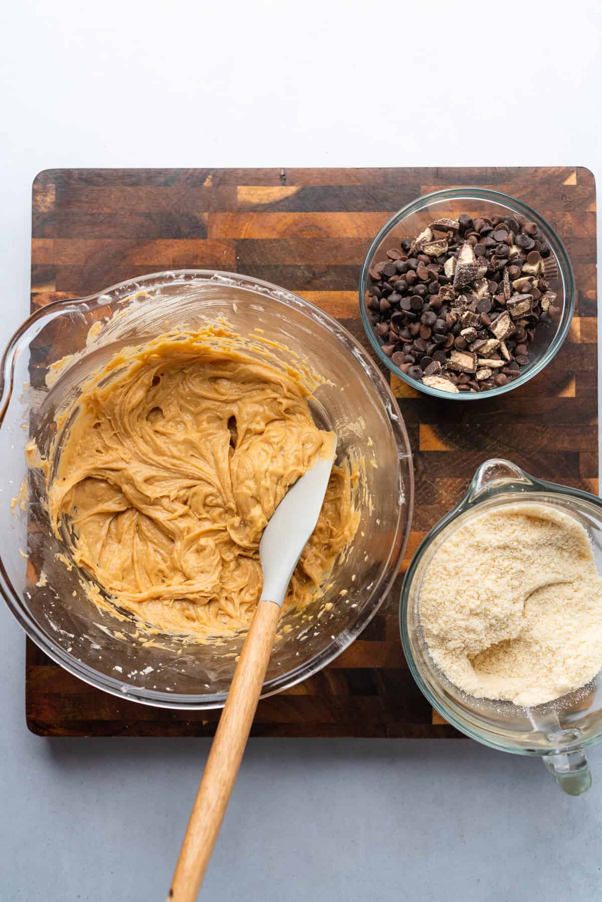 mixed cookie batter in a bowl, with a bowl of chocolate chips and peanut butter cups, and a bowl of almond flour ingredients to make keto peanut butter chocolate chip cookies 