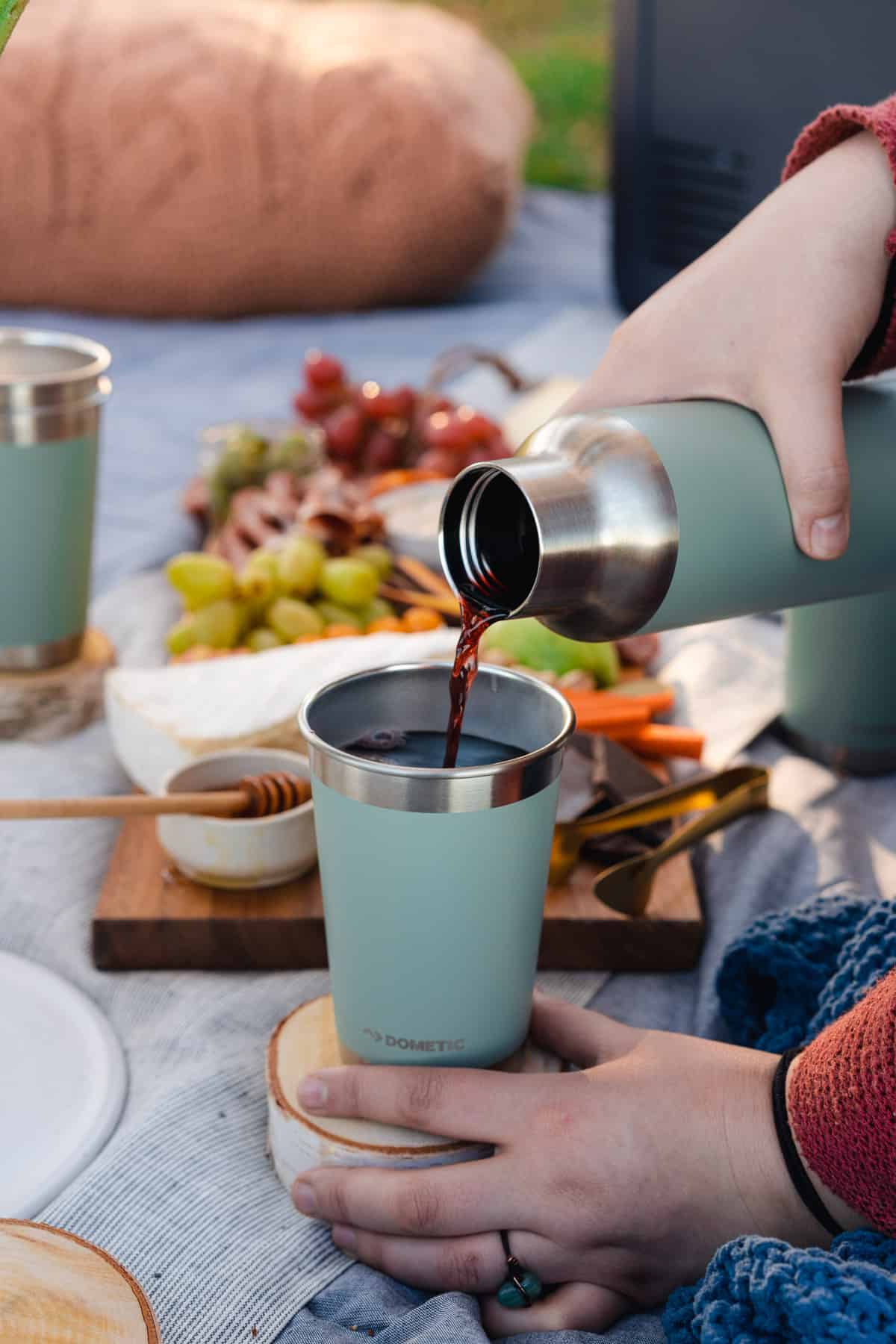 pouring mulled win into a dometic cup with a lovely charcuterie board for a picnic date in the background