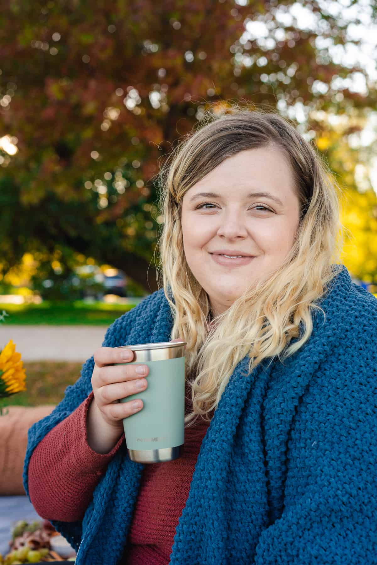 women in a fall setting with a cozy sweater, a blanket wrapped around her, and a tumbler in hand sipping a warm drink