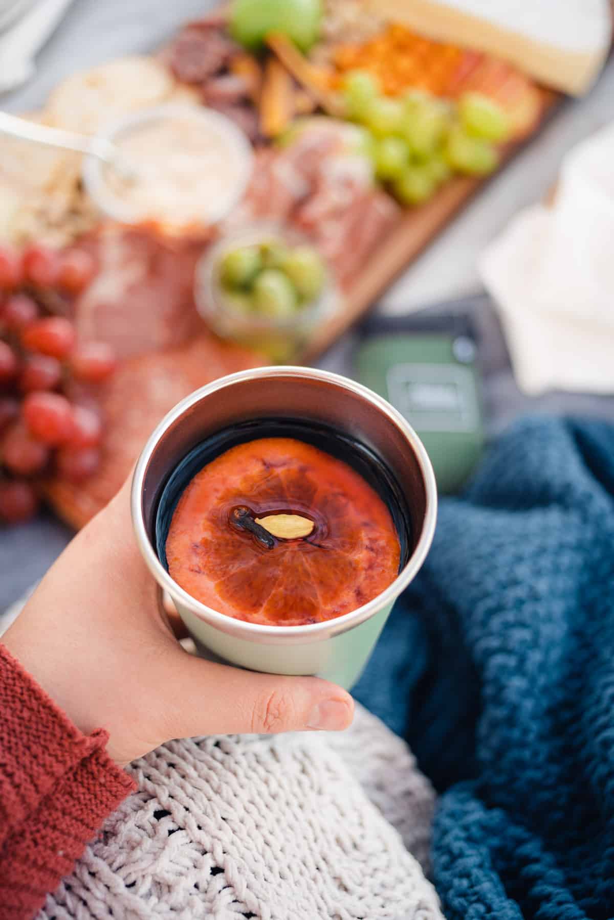 handheld thermal cup with mulled wine over a lovely picnic scene with a snack board