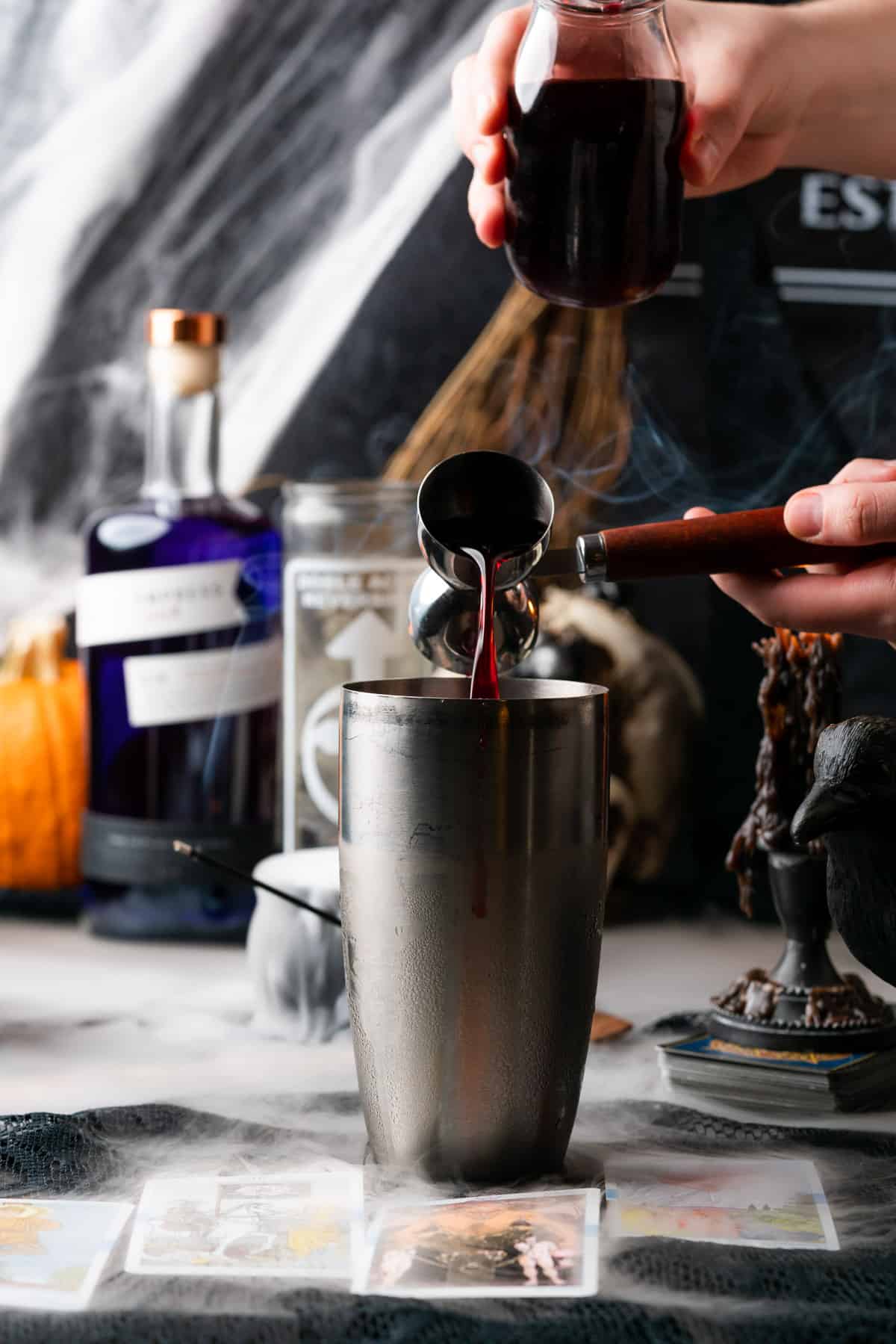 pouring in homemade grenadine into a cocktail shaker with a spooky halloween scene in the background
