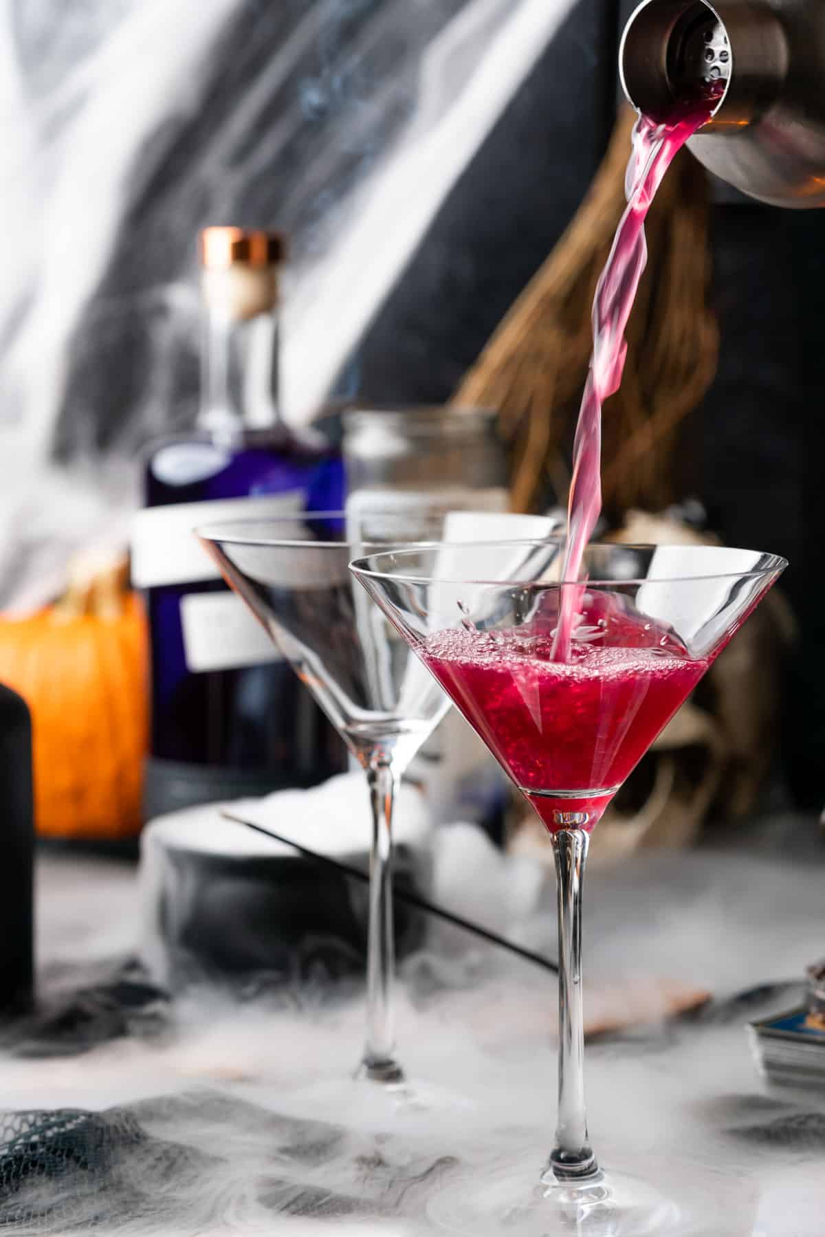 stream of purple cocktail liquid into a martini glass with pruple gin and a pumpkin in the background with dry ice in a cauldron 