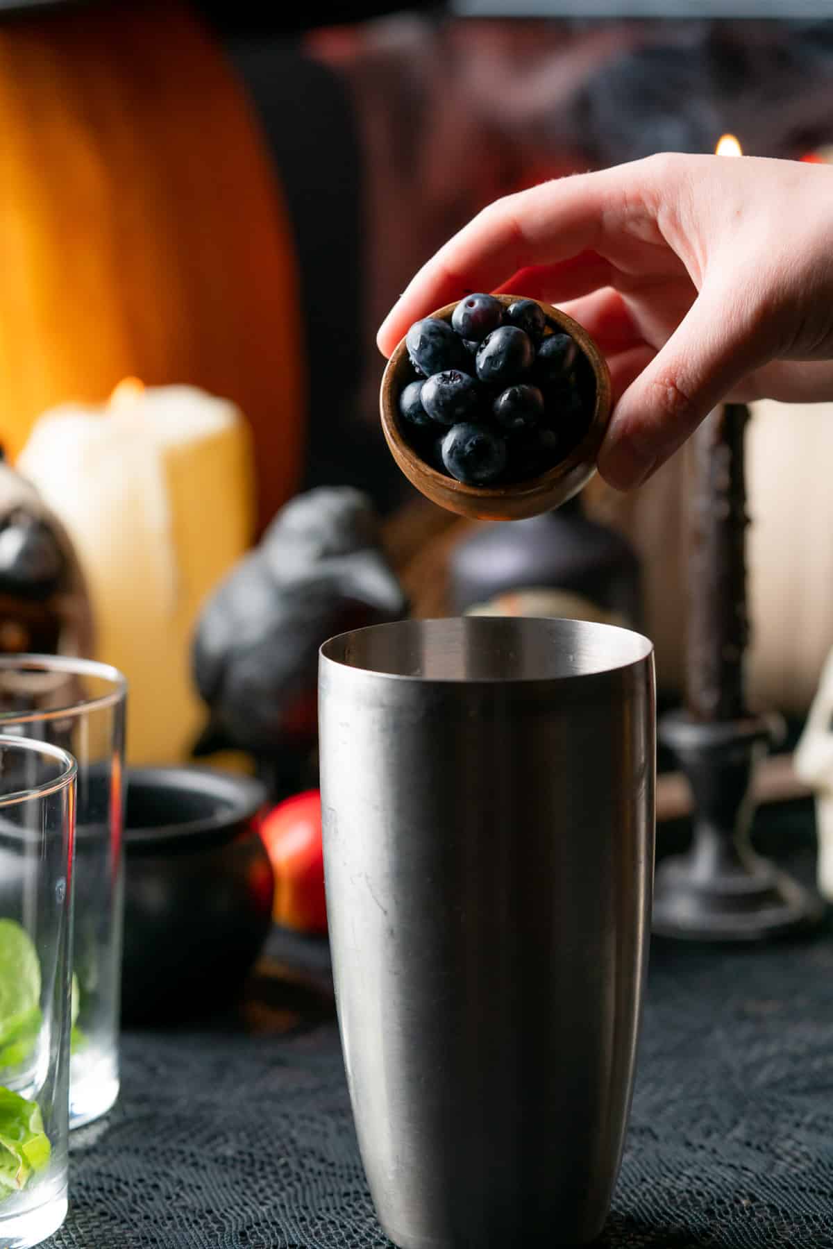 hand pouring blueberries overtop of a cocktail shaker with a spooky halloween backdrop