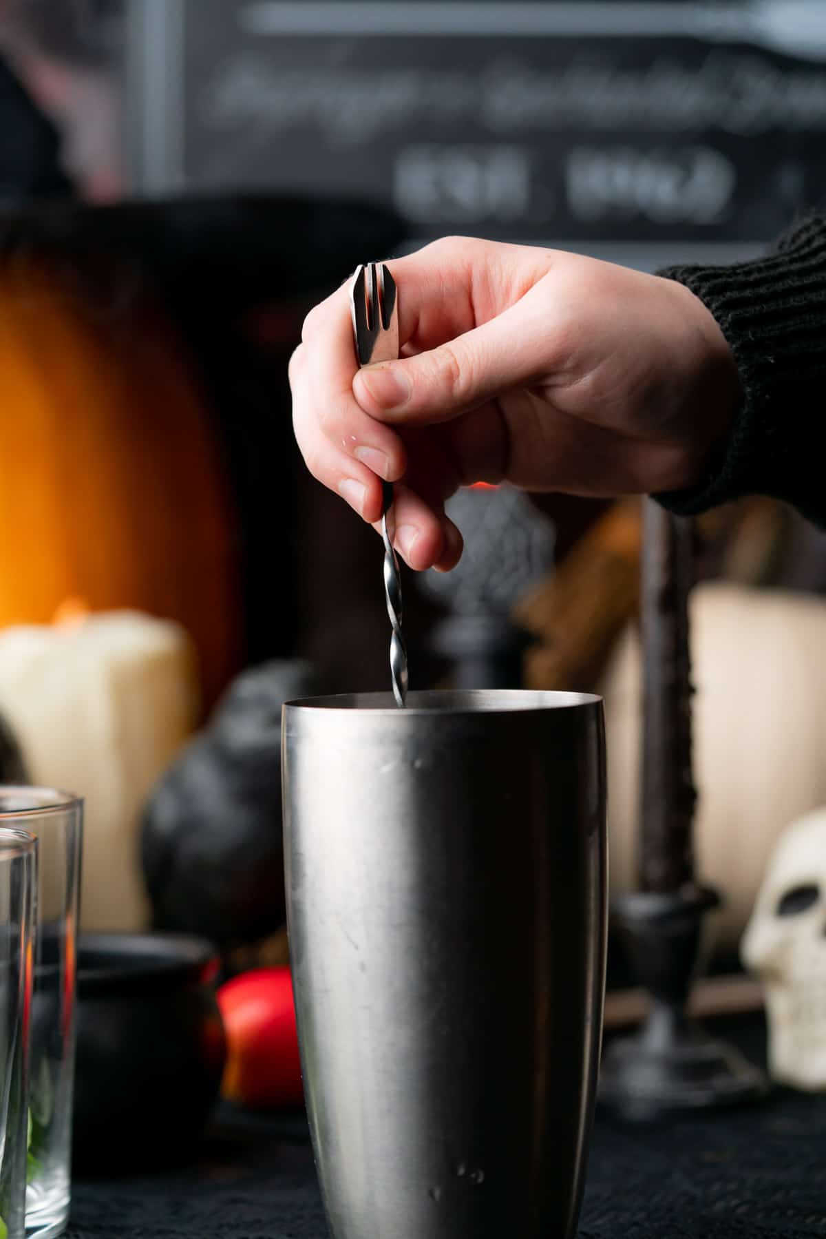 hand stirring a cocktail in a cocktail shaker with a bar spoon with a spooky backdrop