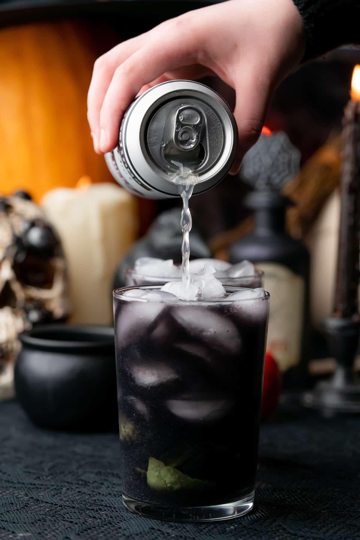 hand pouring ginger beer into a spooky black cocktail in front of a Halloween scene
