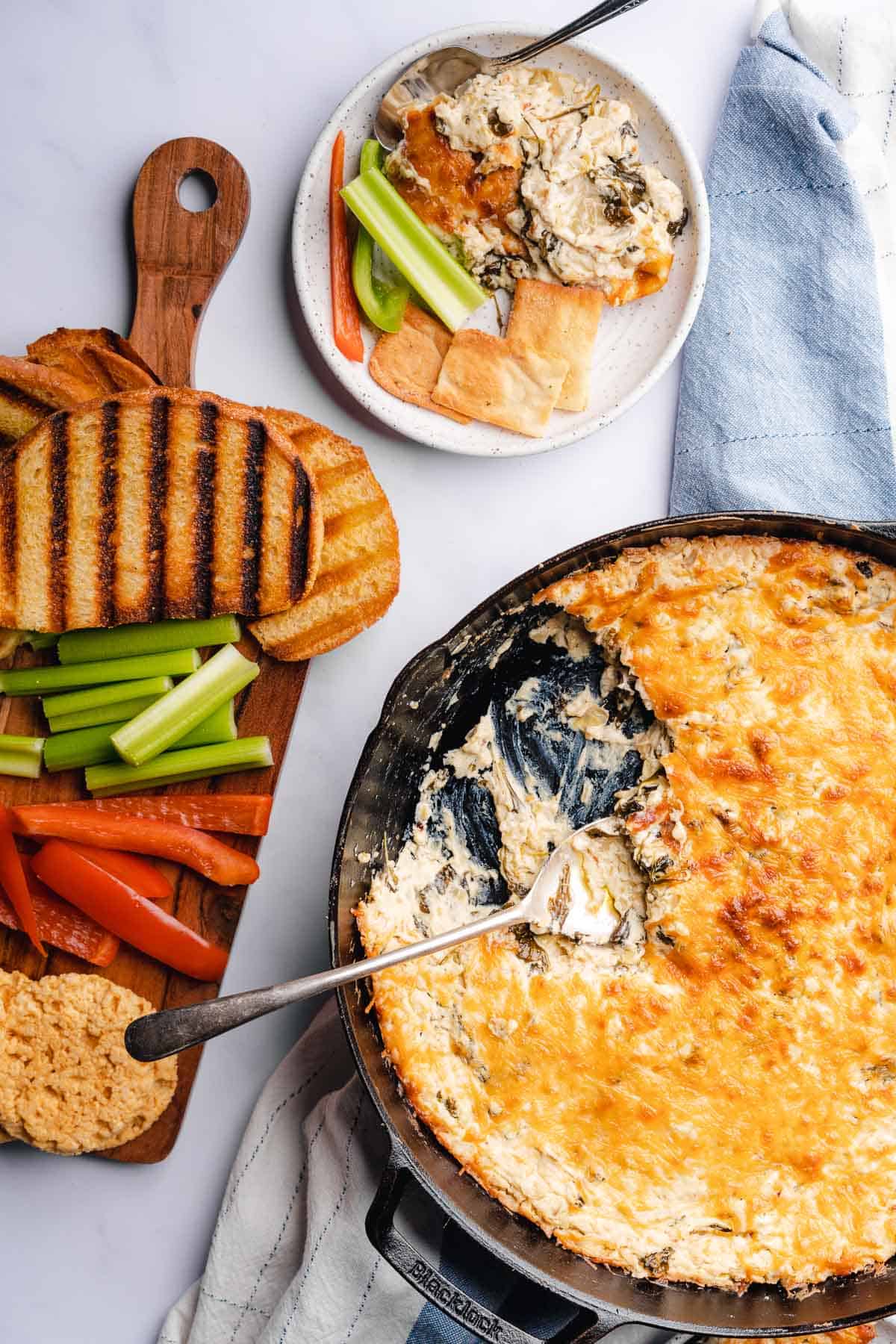 keto spinach artichoke dip in a cast iron skillet, with a large portion taken out of the side , and a plate of grilled sourdough, and vegetables close by