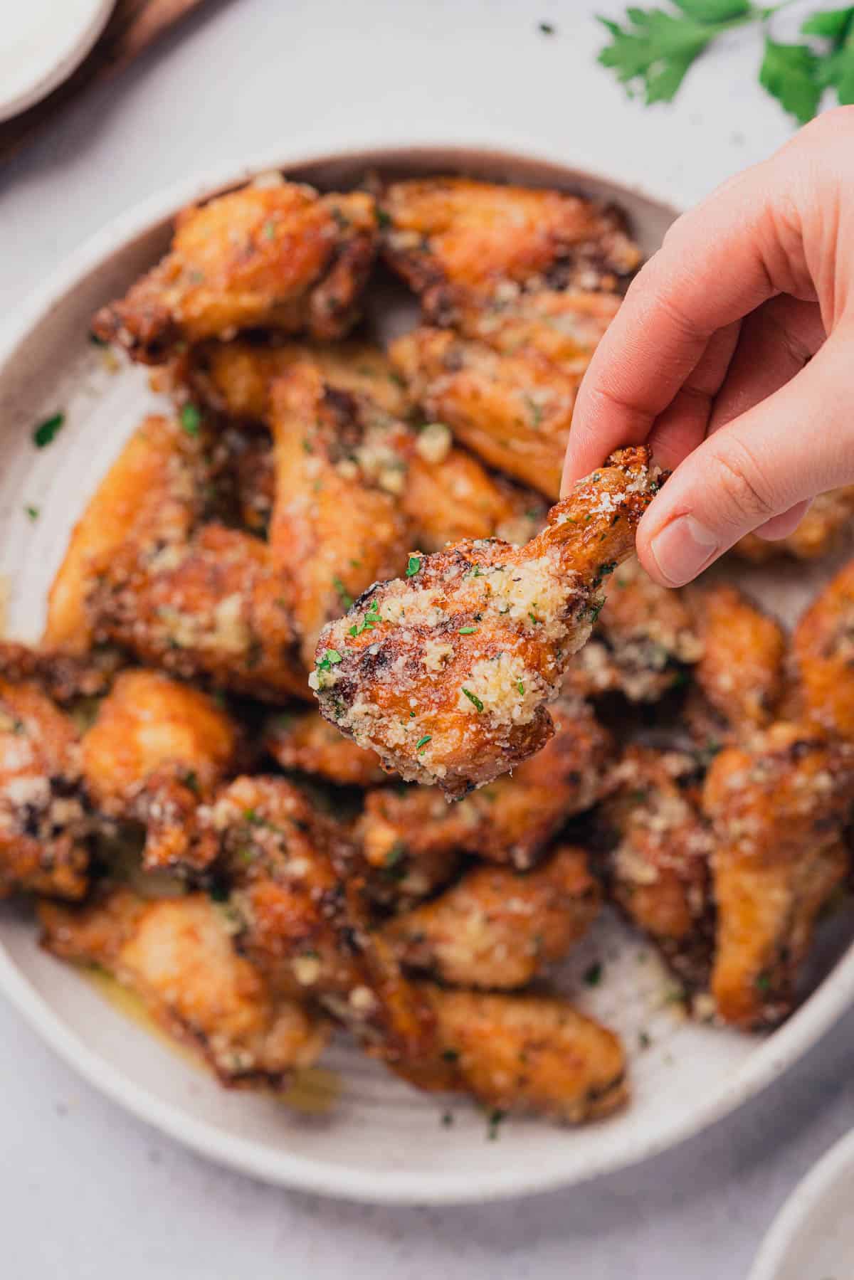 hand holding an air fryer garlic parmesan wing above a full plate of them.