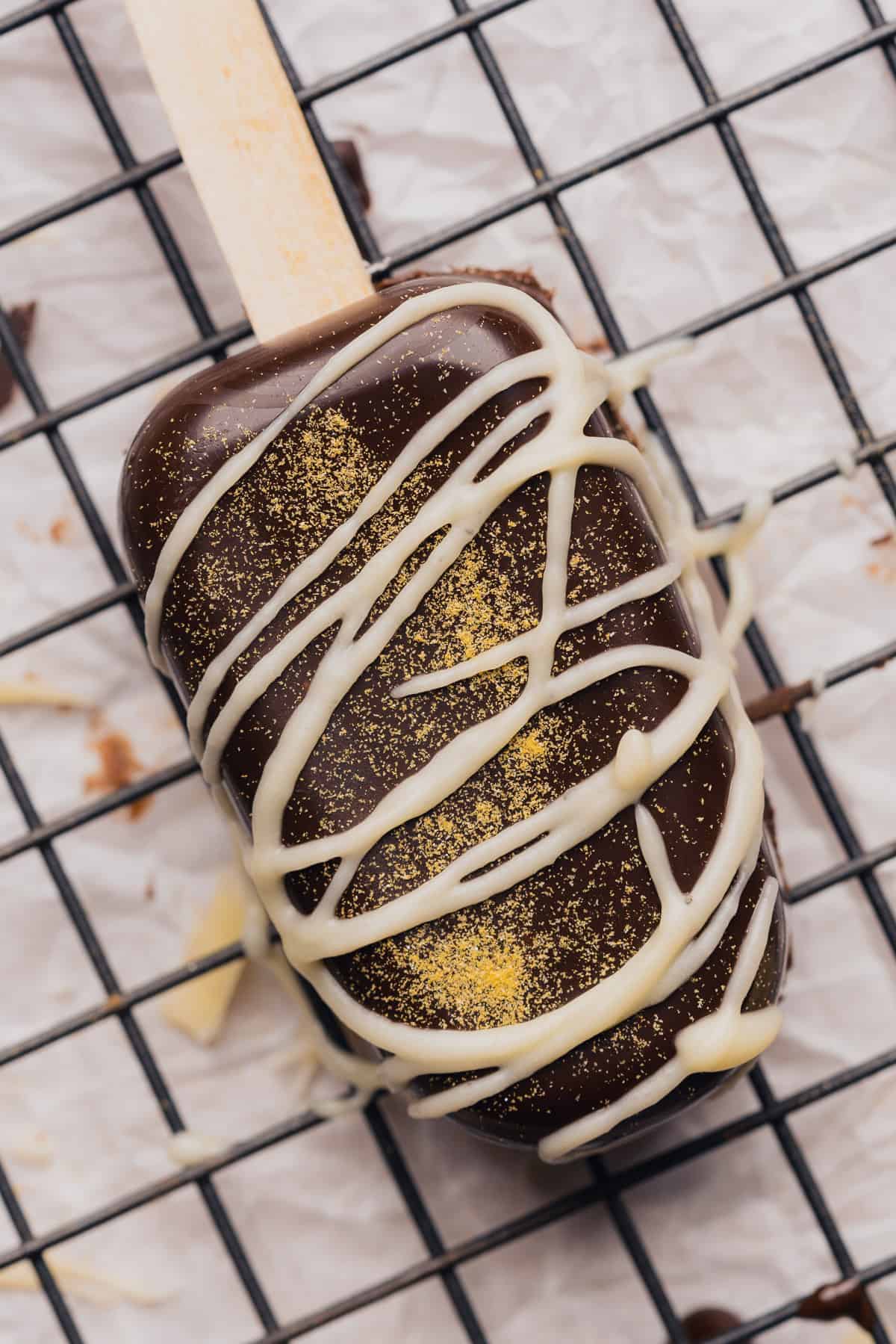 beautiful cakesicle on a black metal cooling rack with a dark chocolate base, gold glitter and white chocolate drizzle 