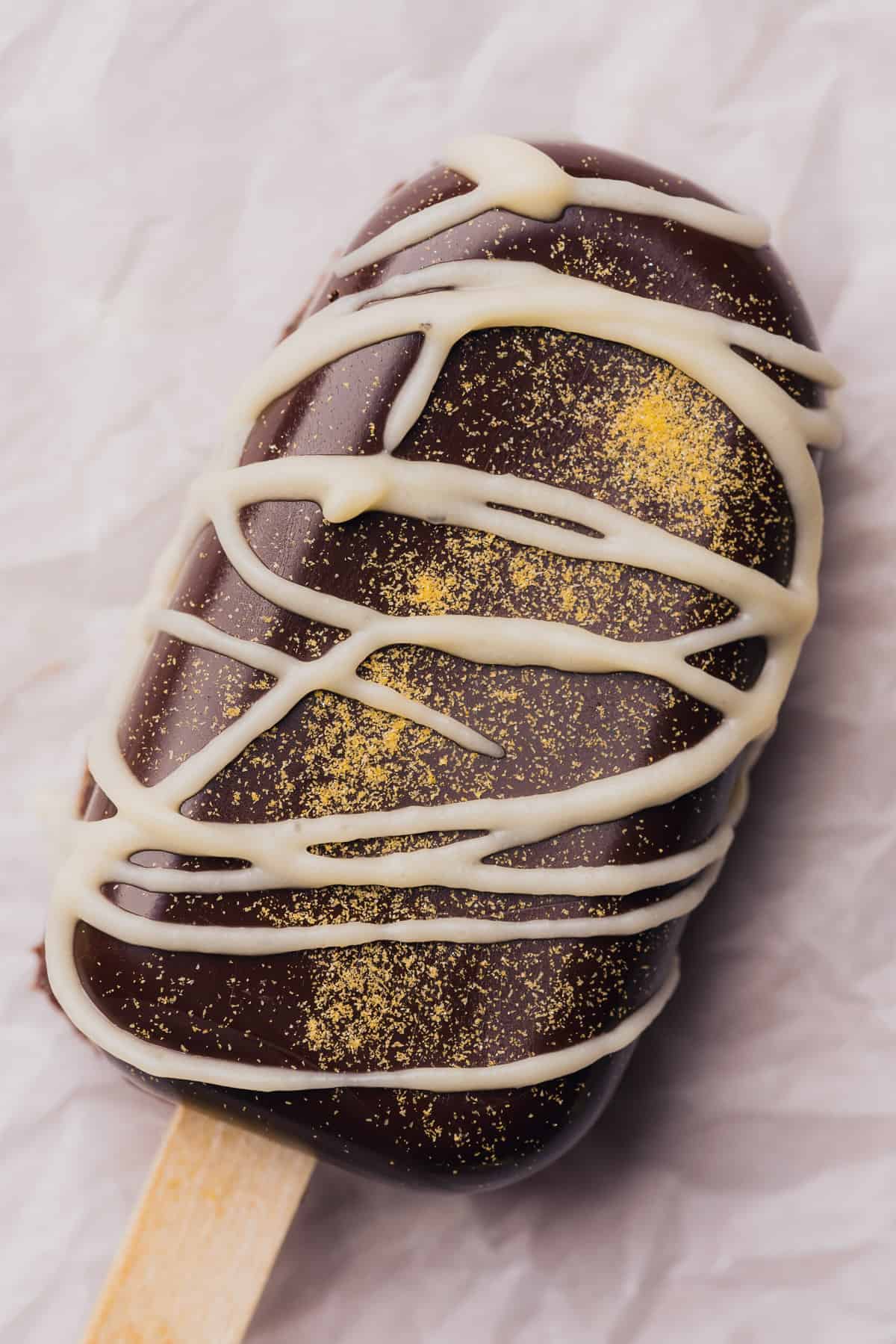 stunning close up shot of a dark chocolate cakesicle with white chocolate drizzle and gold glitter 