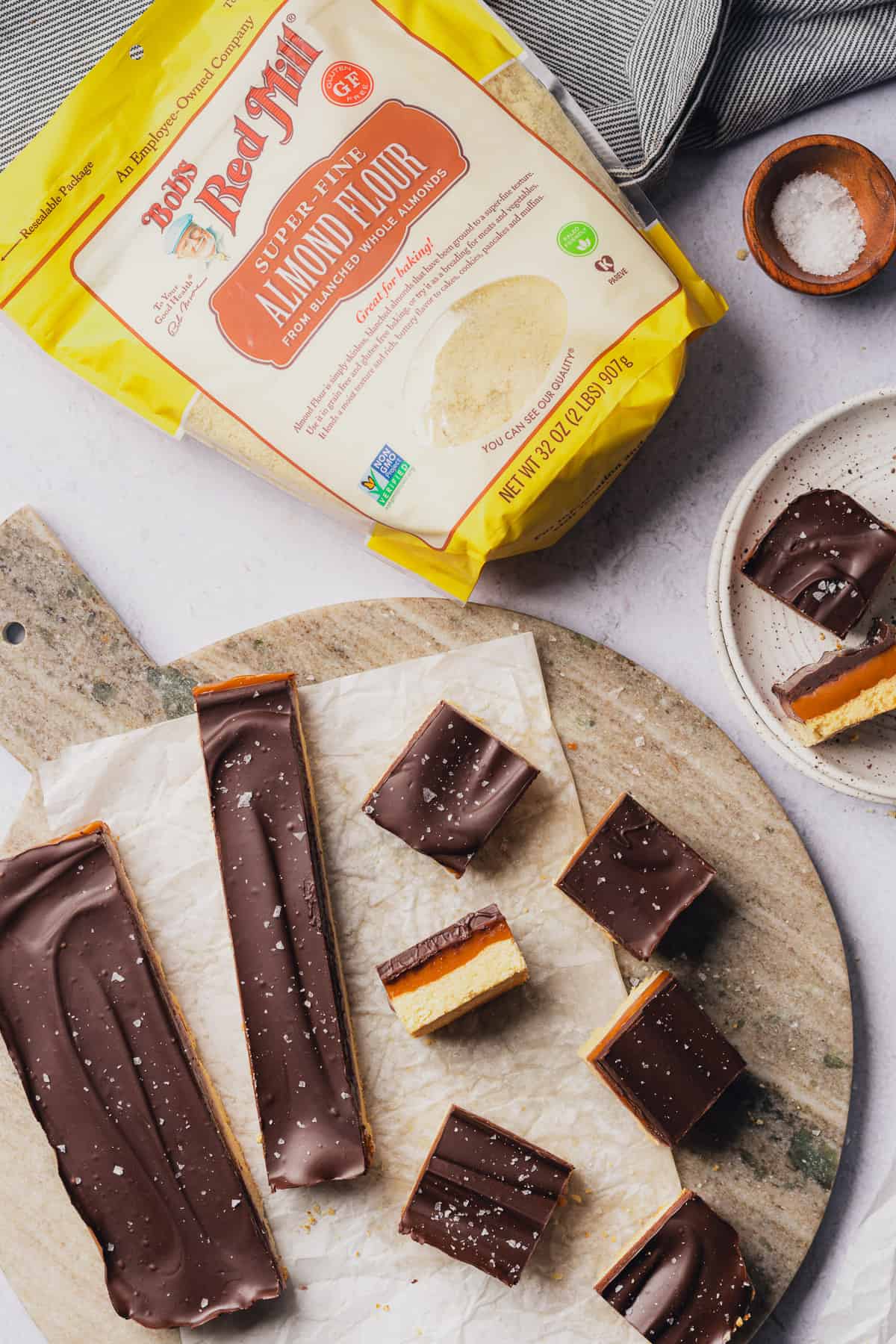super pretty flat lay scene of keto caramel slice bars with  chocolate topping, sea salt and bob's red mill almond flour nearby 