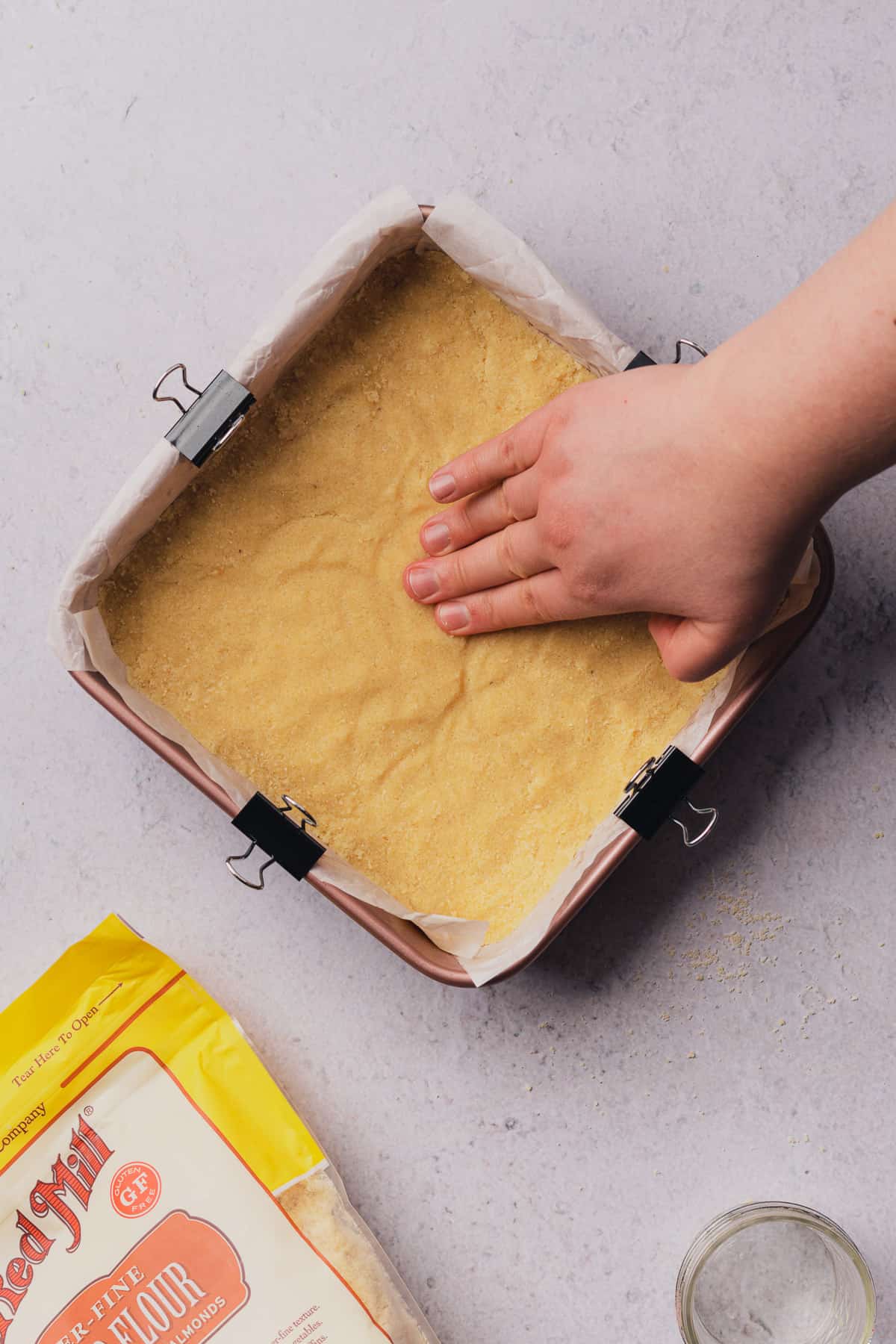 hands pressing down on raw shortbread crust in a 8x8 baking pan with parchment