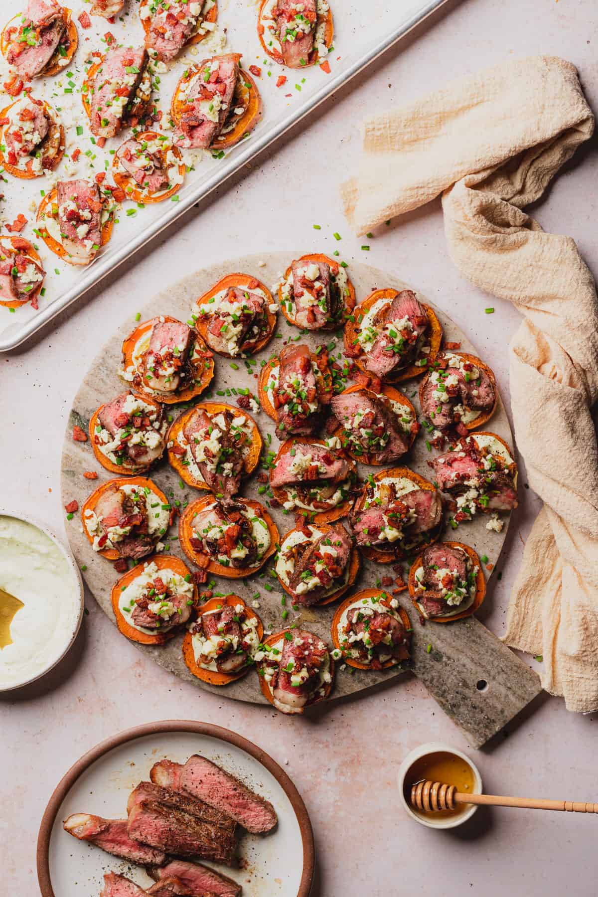 steak appetizer with sweet potato bacon and blue cheese on a grey marble round with a sheet pan full of steak bites
