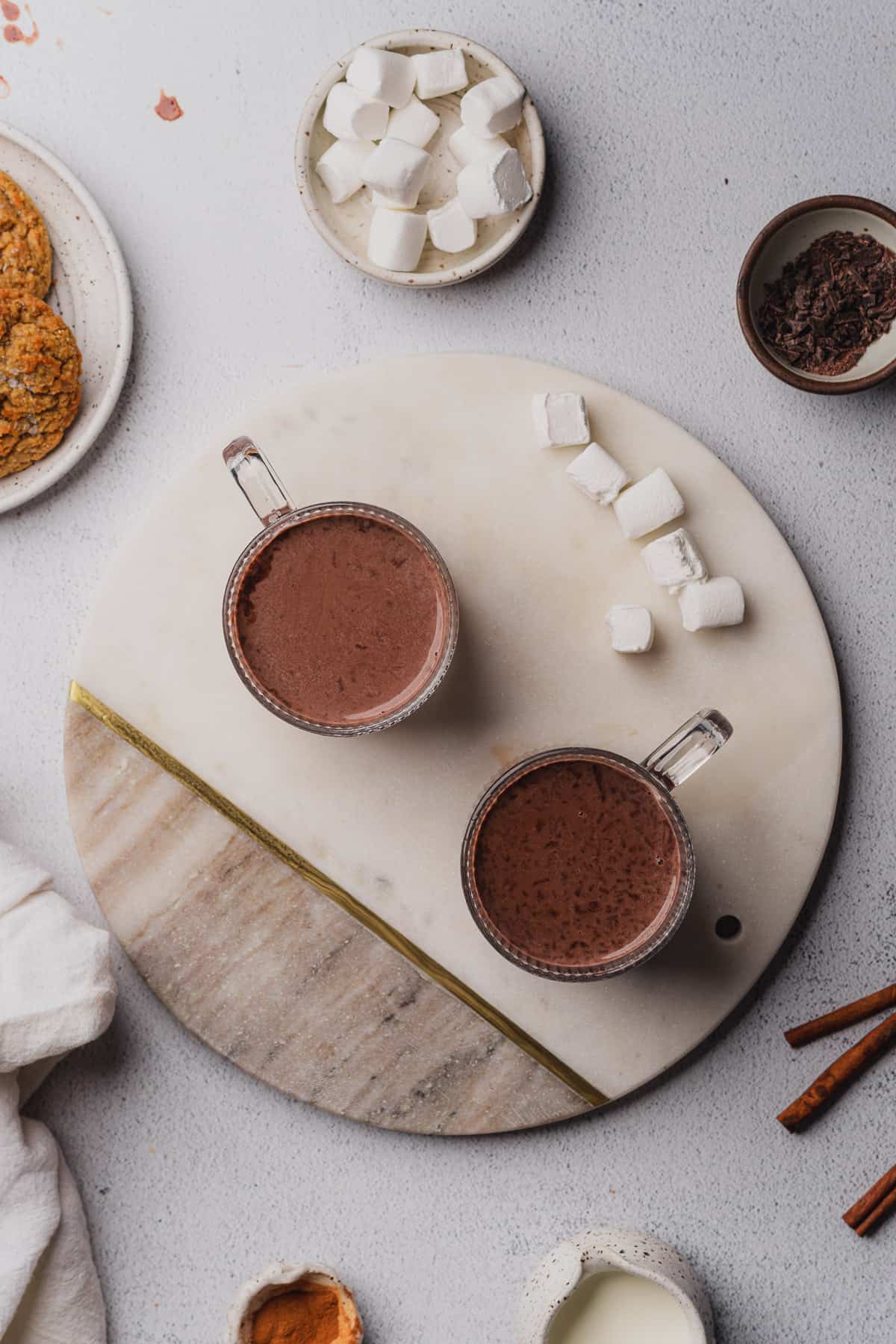 hot chocolate in glass mugs surrounded by cinnamon sticks, marshmallows and cookies