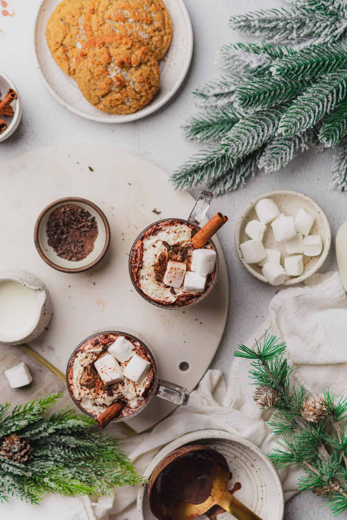 flat lay shot of instant pot hot chocolate topped with whipped cream, marshmallows, cinnamon sticks, chopped chocolate, with garland and cookies nearby 