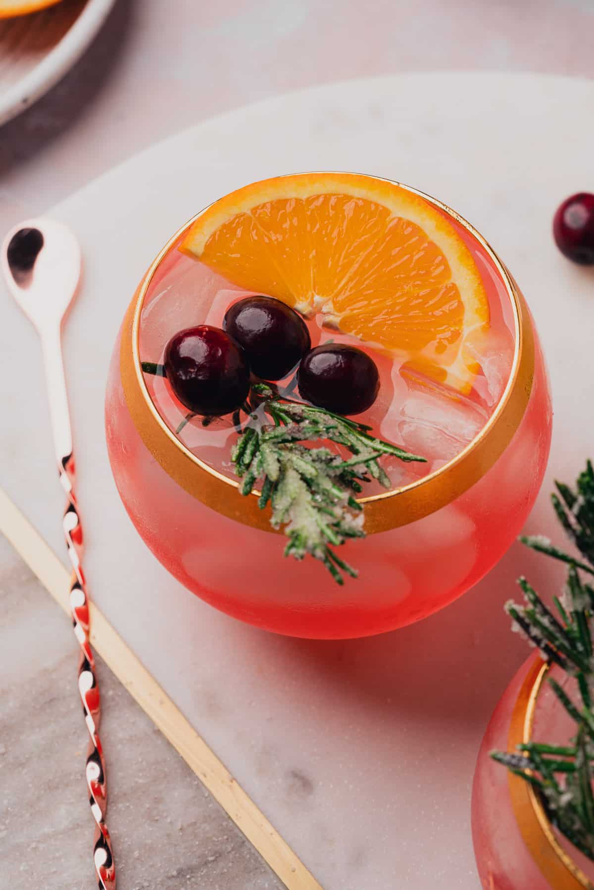 close up shot of a cranberry prosecco margarita on a marble white and grey board with gold, in a round goblet with a gold rim, and a bar spoon nearby