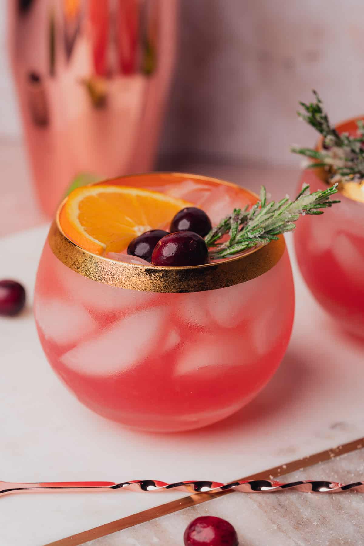 side shot of two round goblet glasses with an orange slice, cranberries, and rosemary on top 