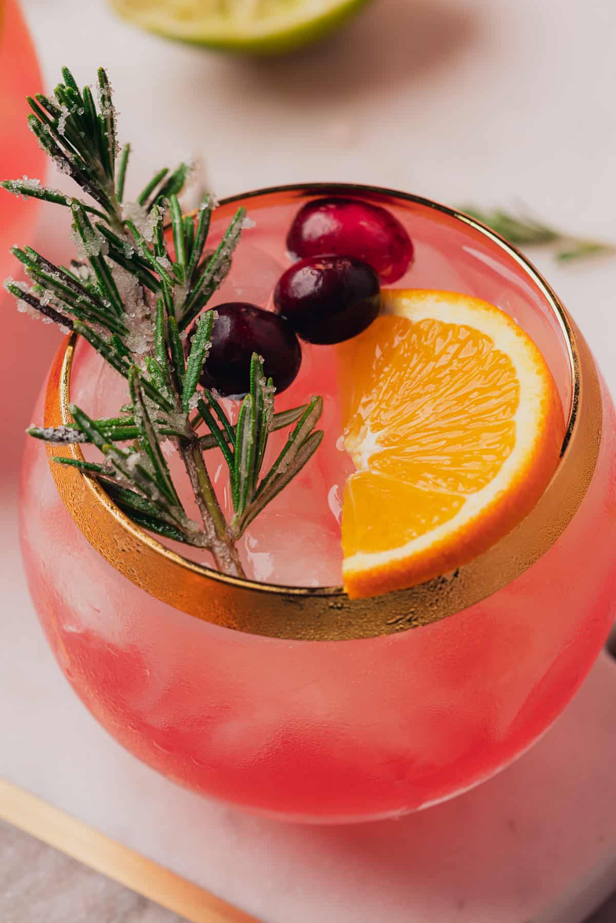 prosecco margarita with cranberries, an orange slice and a sugared piece of rosemary in a goblet with a gold rim