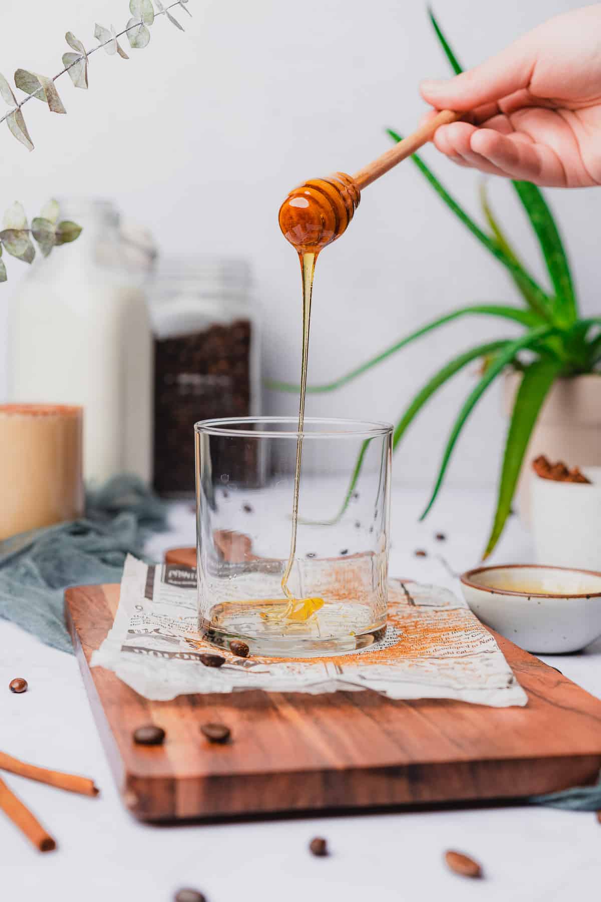 hand pouring honey into a glass surrounded by a plant and coffee making ingredients