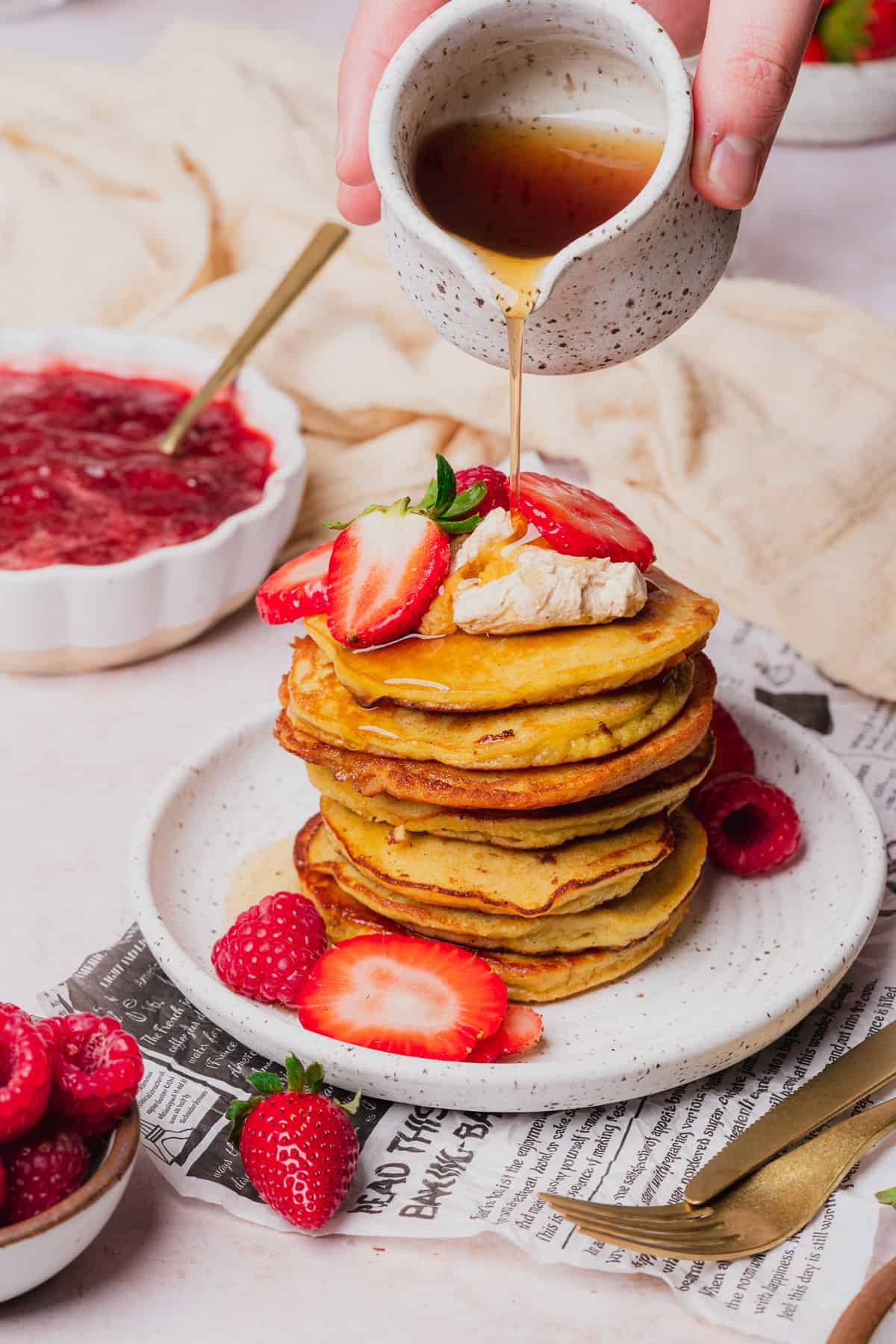 pouring maple syrup over top of a stack of keto coconut flour pancakes topped with whipped butter, fresh raspberries and strawberries with strawberry jam in the background