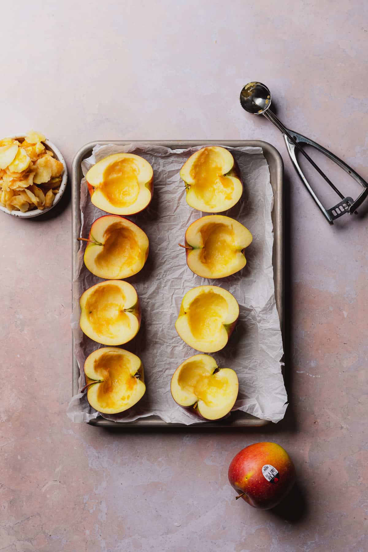 apples on a parchment lined baking sheet, with a cookie scoop and apple flesh scooped out to reveal a hollow apple shell. 