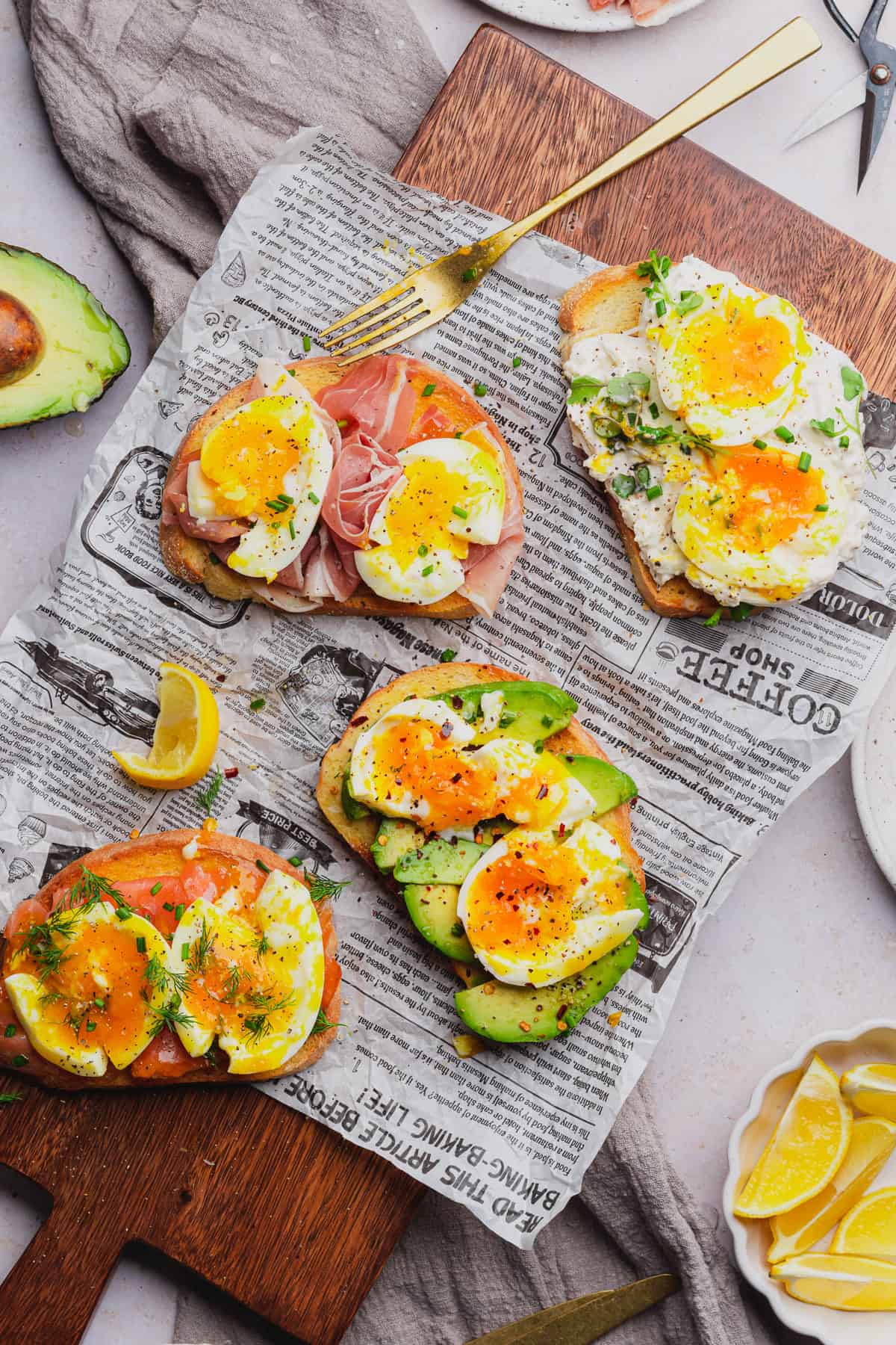 beautiful smashed eggs with runny yolks on toast with prosciutto, avocado, smoked salmon, fresh herbs and burrata cheese on a newspaper