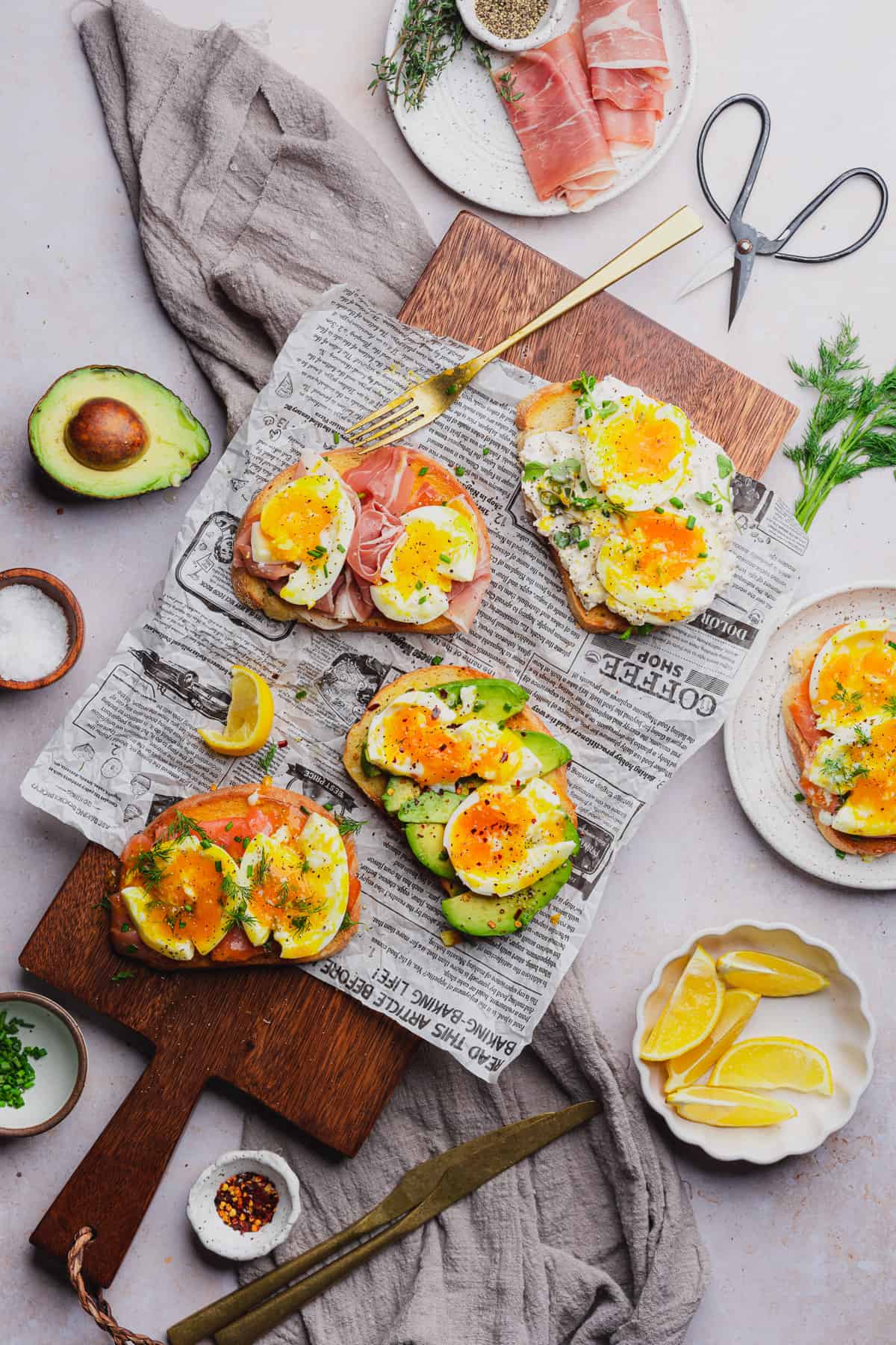 flat lay of 5 slices of toast with smoked salmon, burrata, avocado, and prosciutto topped with yolky smashed eggs, fresh herbs, and lemon