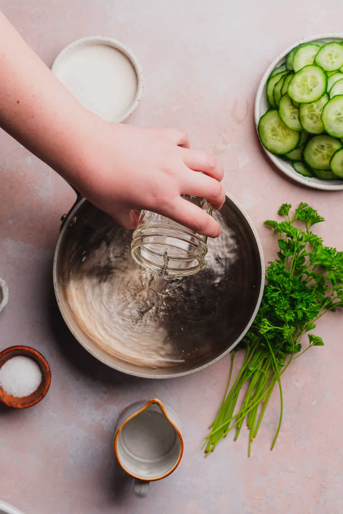 hand pouring vinegar into a saucepan surrounded by cucumbers, seasonings, and parsley.