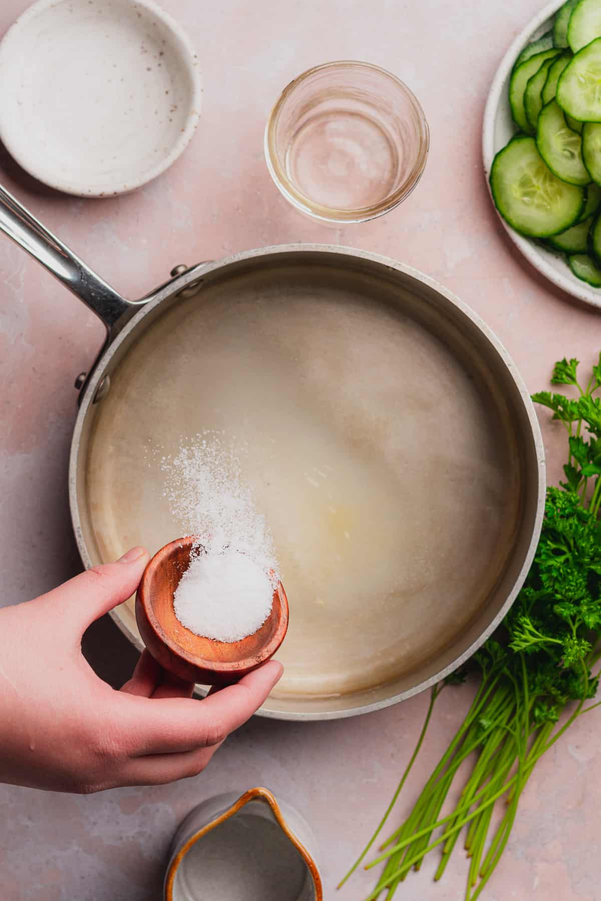 hand pouring salt into a saucepan surrounded by cucumbers, seasonings, and parsley.