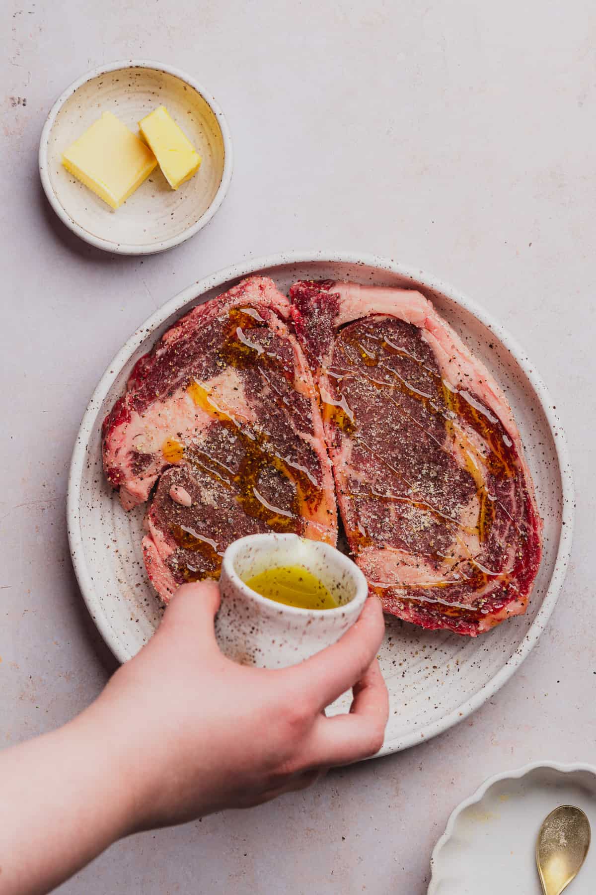 hand pouring olive oil over top of seasoned ribeye steaks
