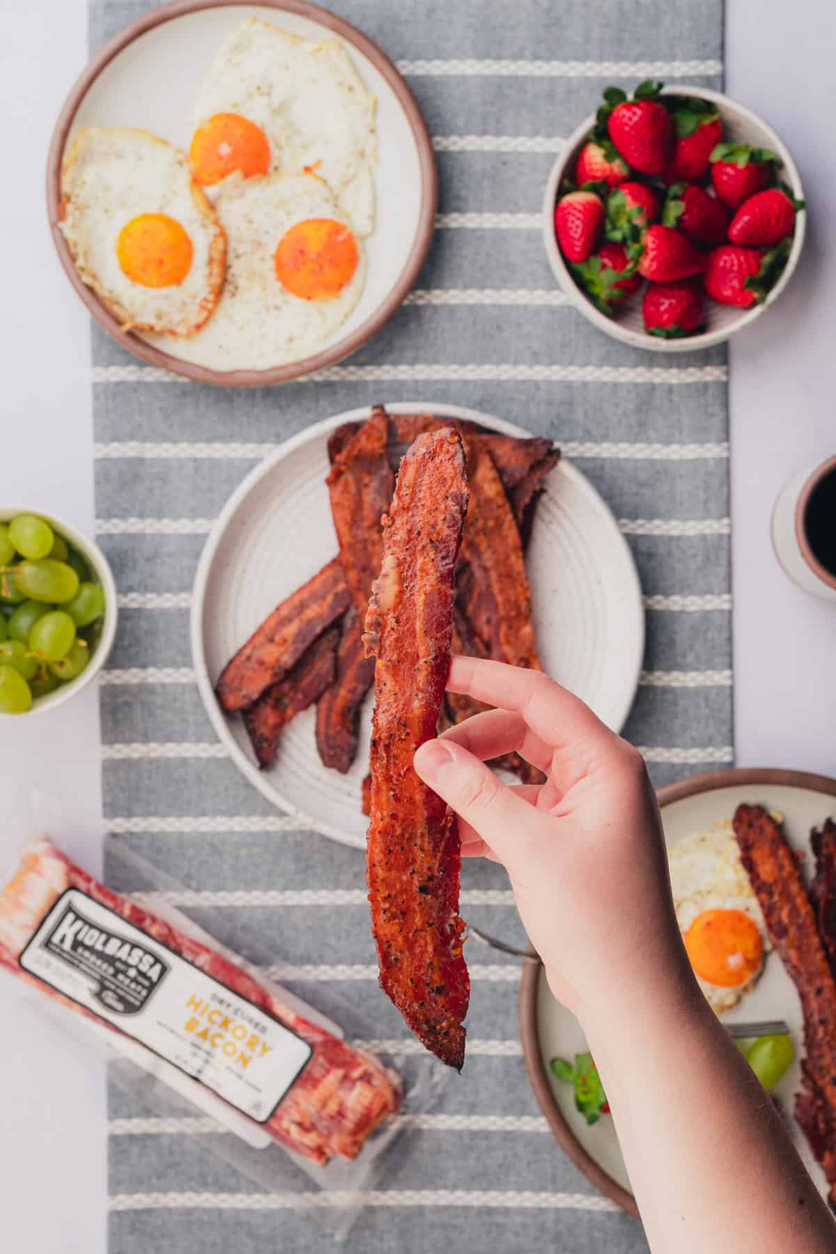 hand holding up a slice of keto candied bacon to the screen with a breakfast scene below