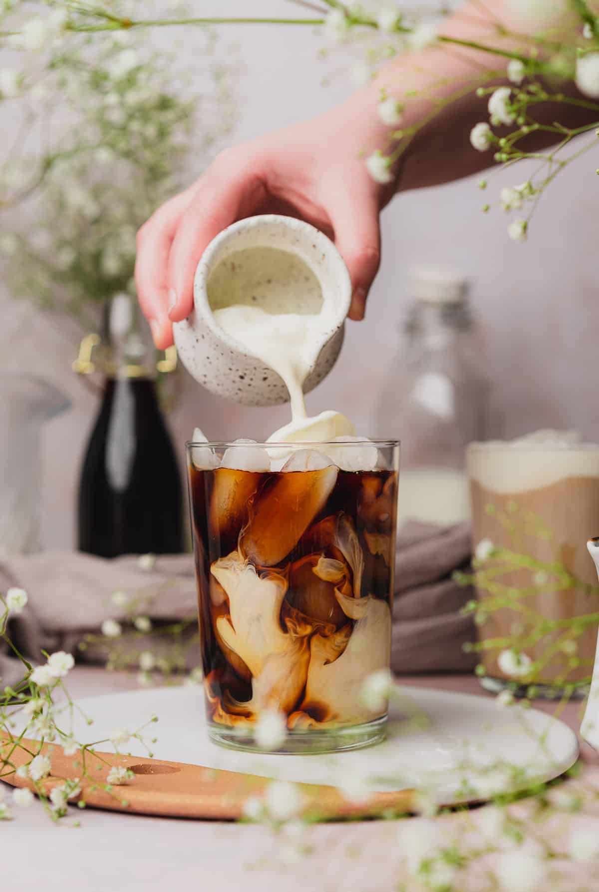 hand pouring sweet cream cold foam on top of iced coffee with baby's breath flowers in the background.
