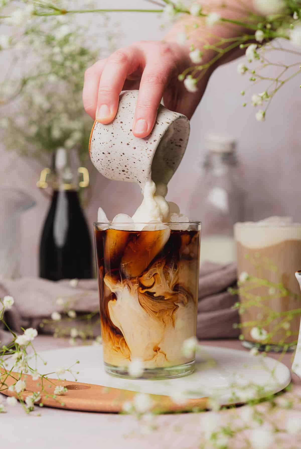 pouring sweet cream cold foam on top of iced coffee with baby's breath flowers in the background and foreground.