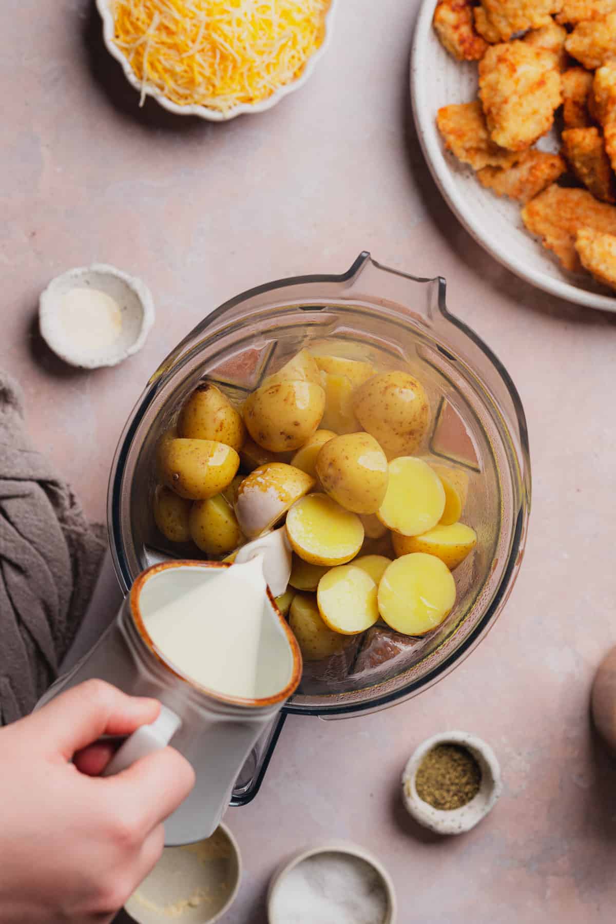hand pouring heavy cream into a blender of cooked potatoes. 